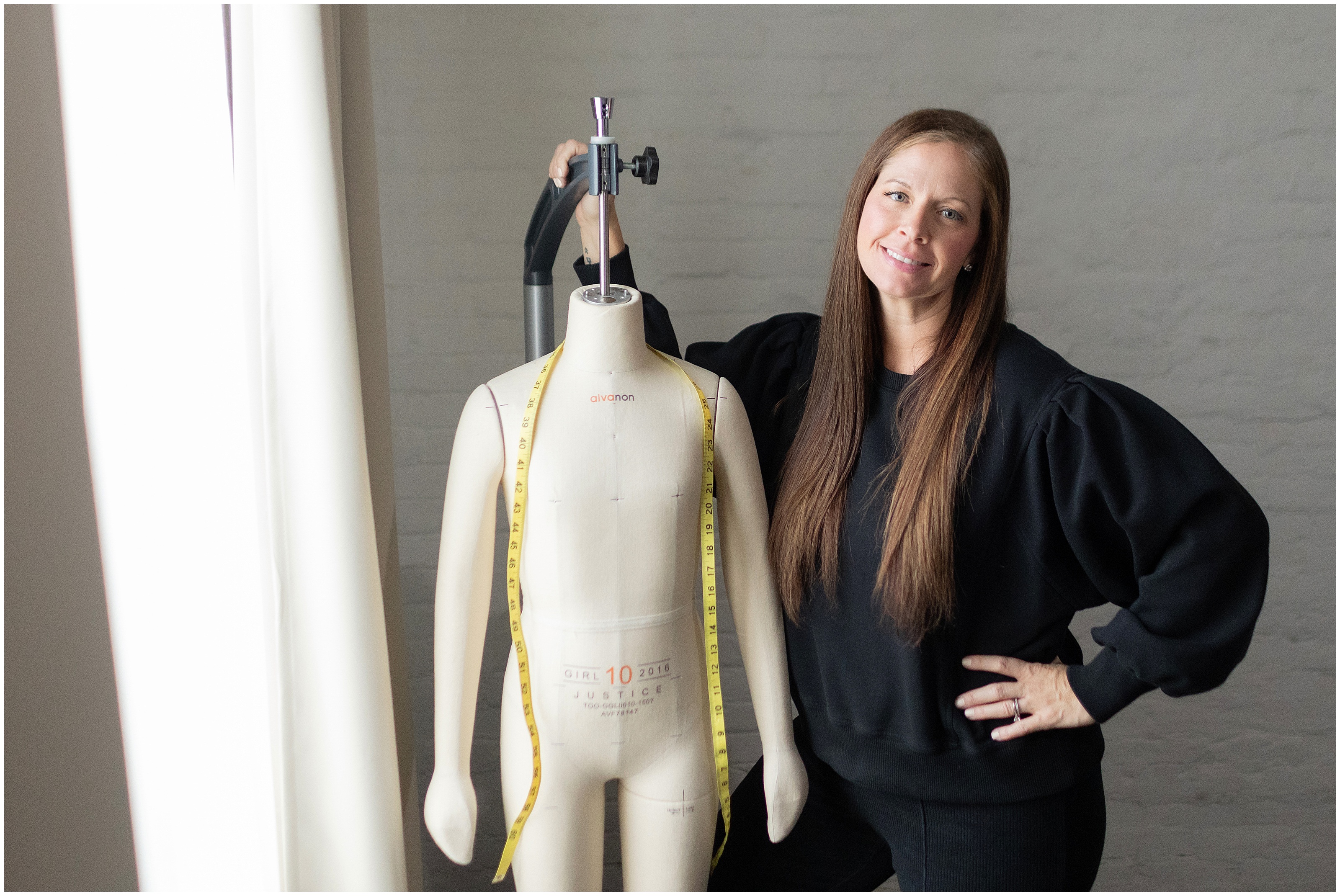 Woman in black smiles beside a dress form with measuring tape in a design studio
