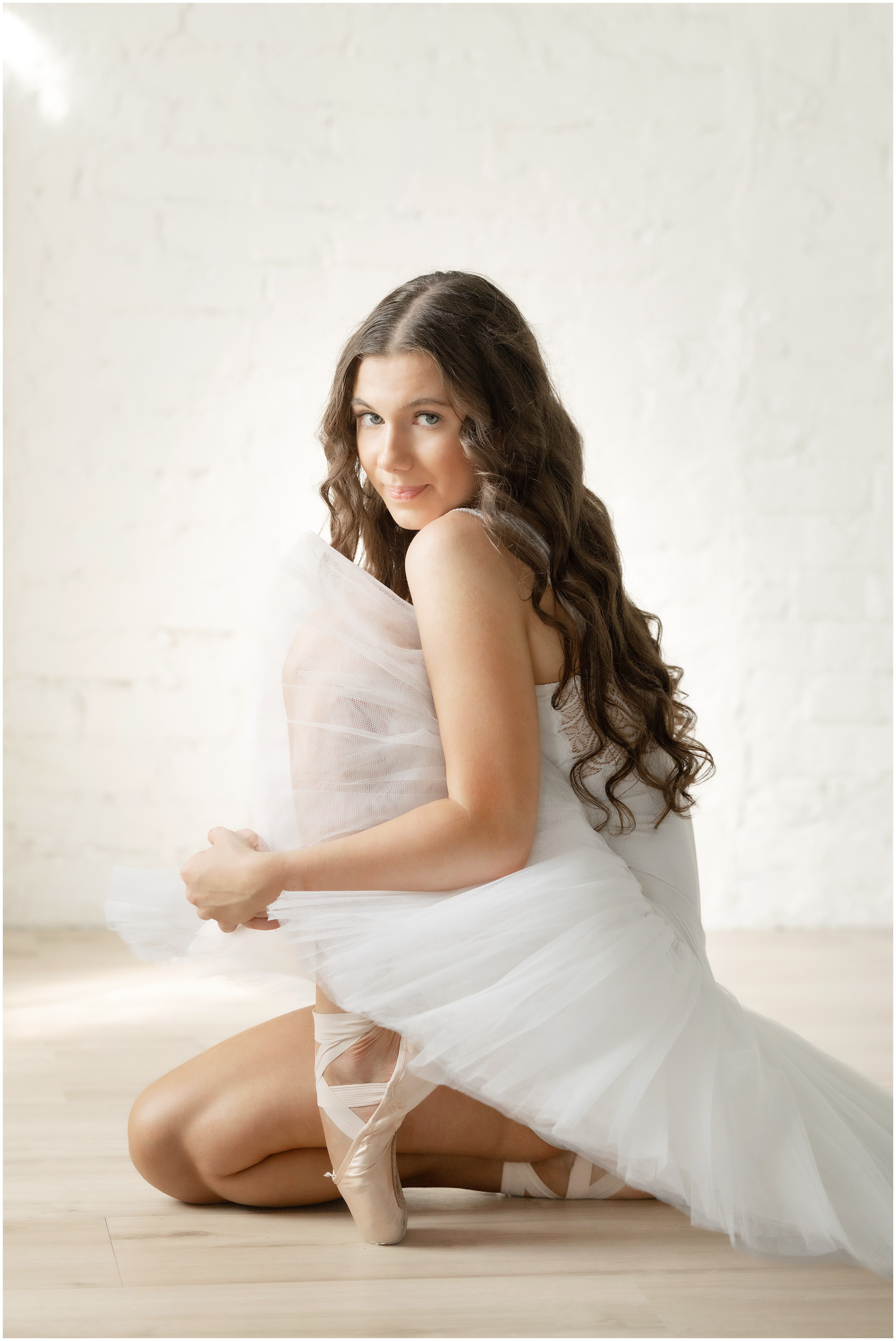 Smiling ballerina in a white tulle dress kneels on a light wood floor against a bright studio backdrop.