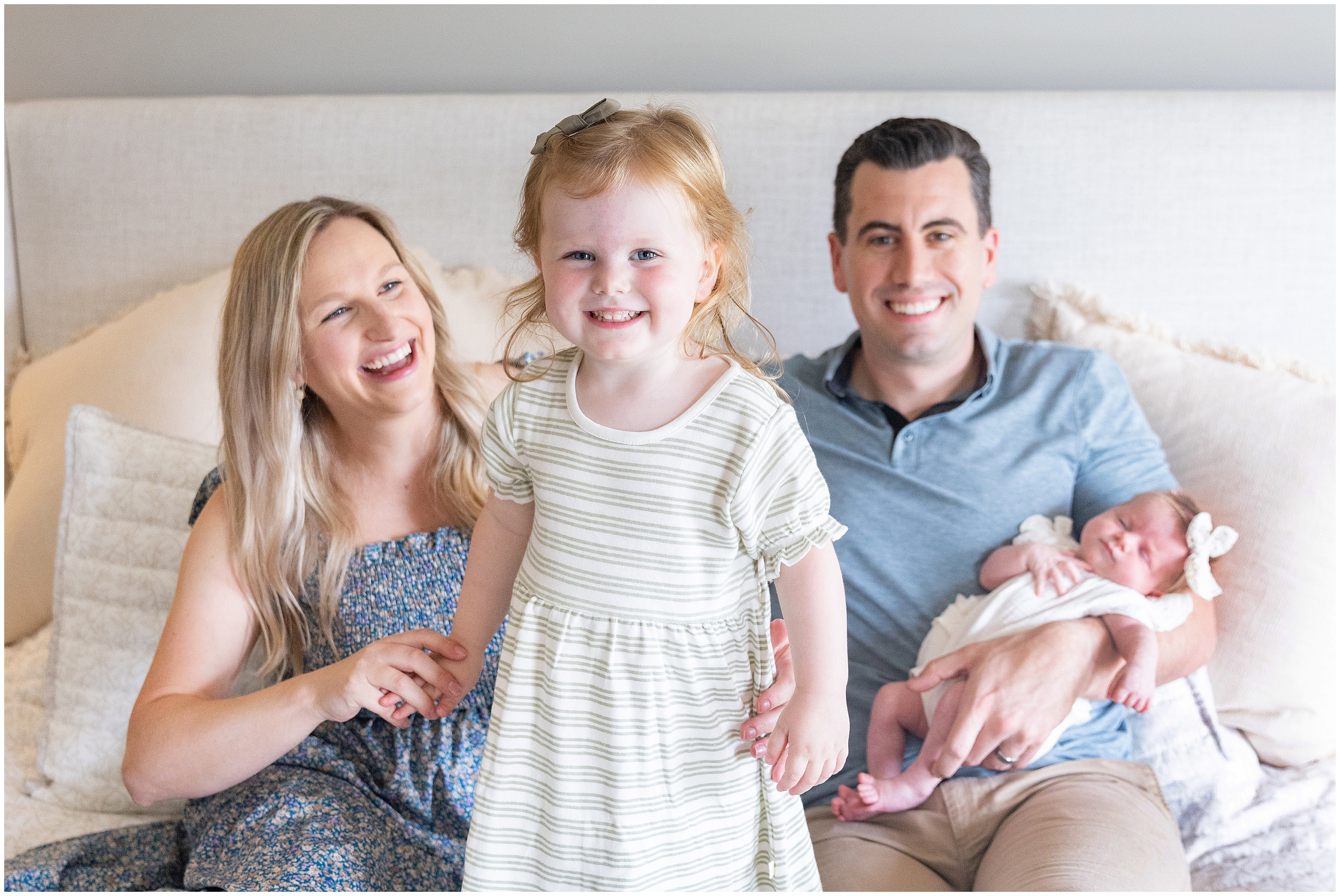 Smiling family of four sitting on a bed, together, parents with toddler and newborn in a bright bedroom with cream pillows and bedding. Columbus Ohio newborn photography