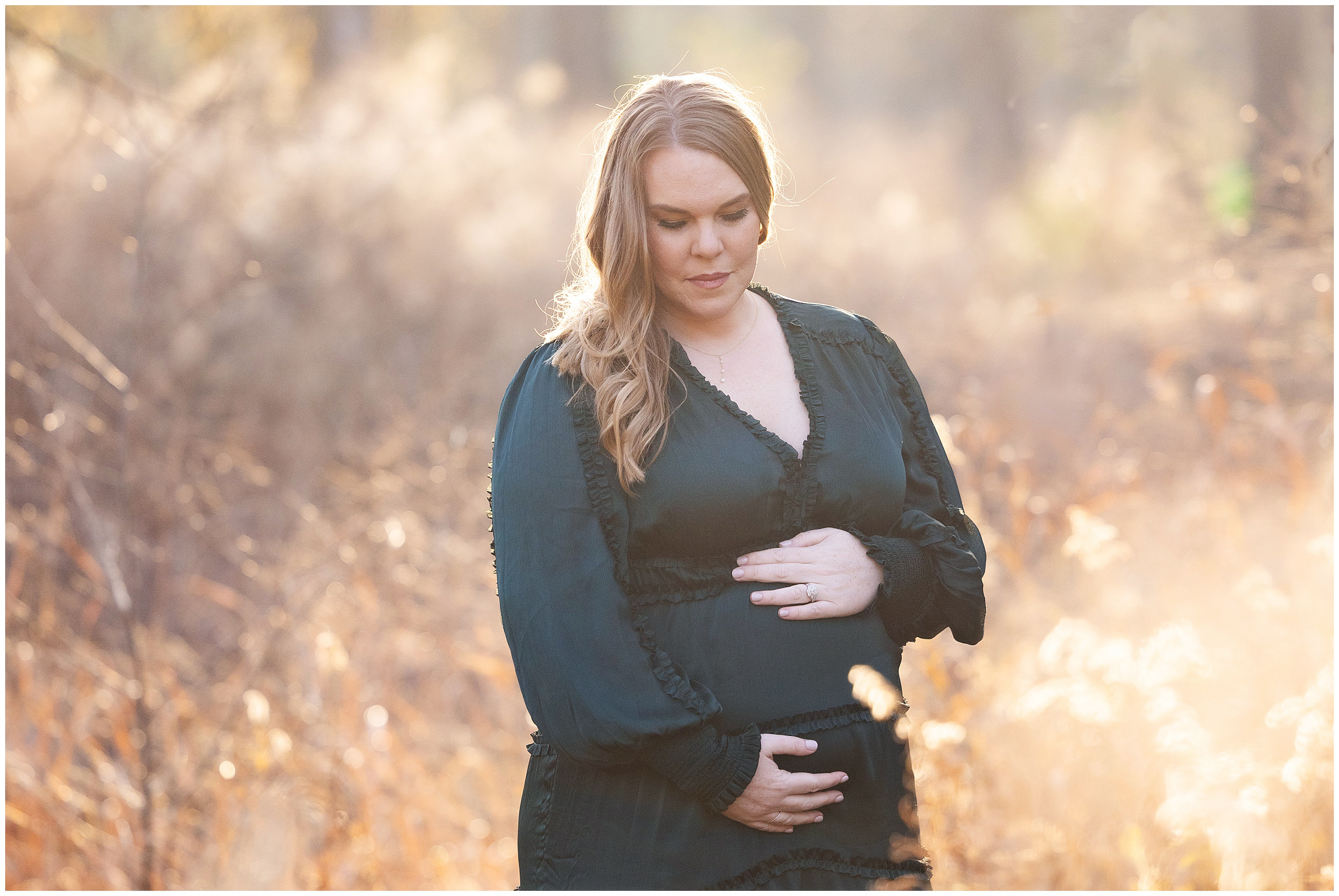 Pregnant woman in a dark green dress, cradling her belly in a sunlit, autumn field. Soft, golden light creates a serene atmosphere.