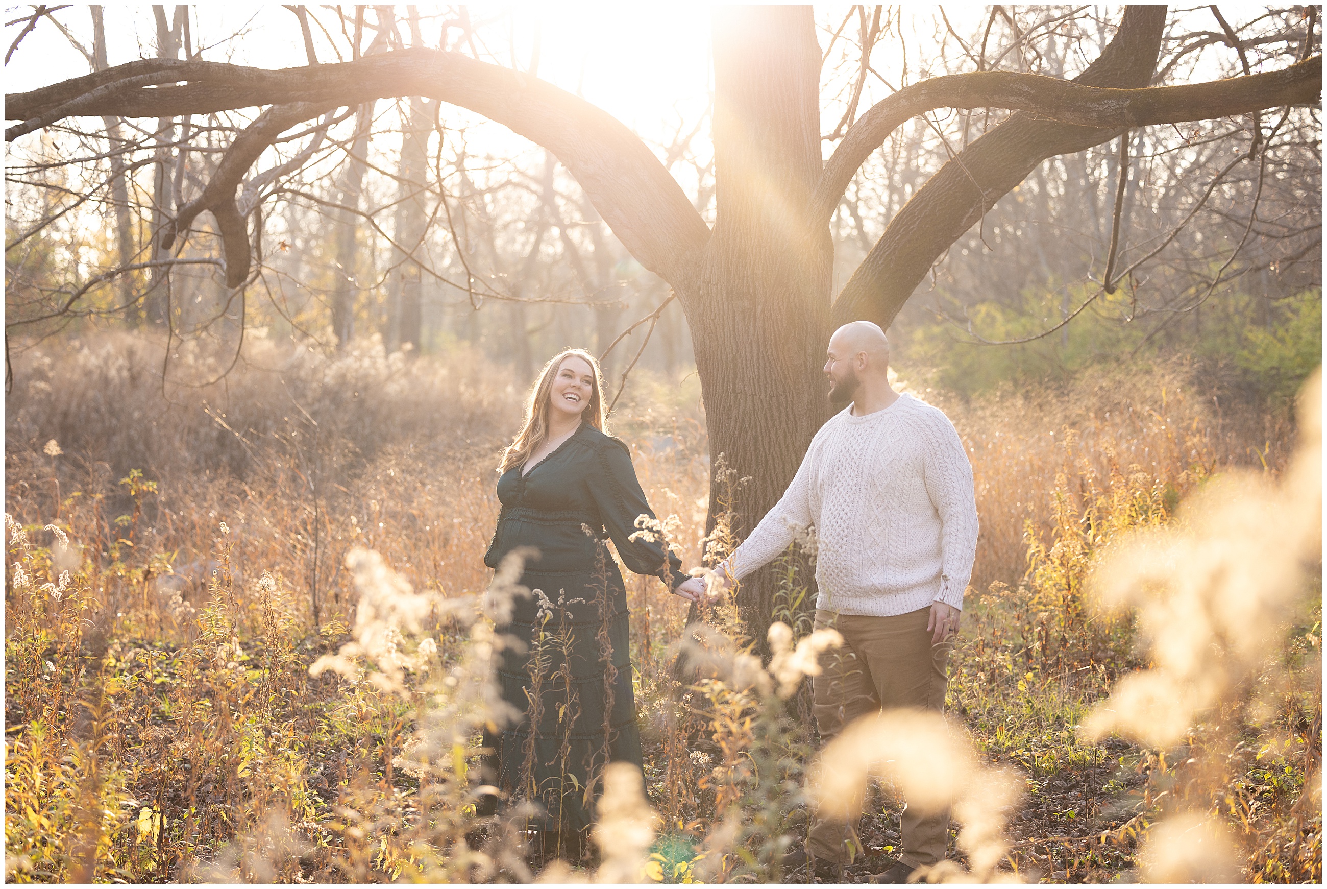 A couple holds hands joyfully in a sunlit meadow, surrounded by tall grass and bare trees, under a warm autumn glow. Columbus, Ohio maternity photographer.