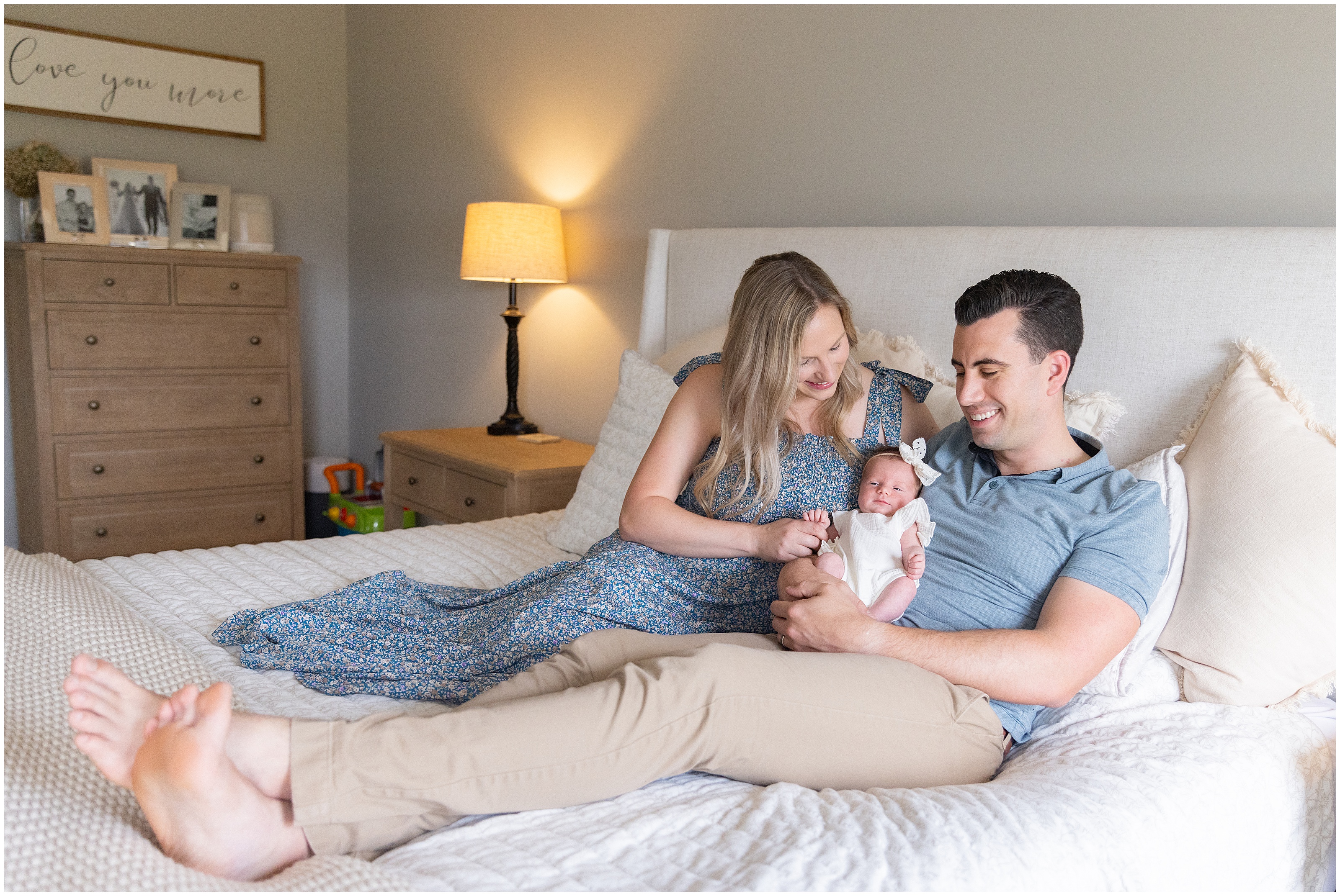 Smiling couple sits on a bed holding their newborn in a cozy bedroom, Smiling family of four sitting on a bed, together, parents with toddler and newborn in a bright bedroom with cream pillows and bedding. Columbus Ohio newborn photography