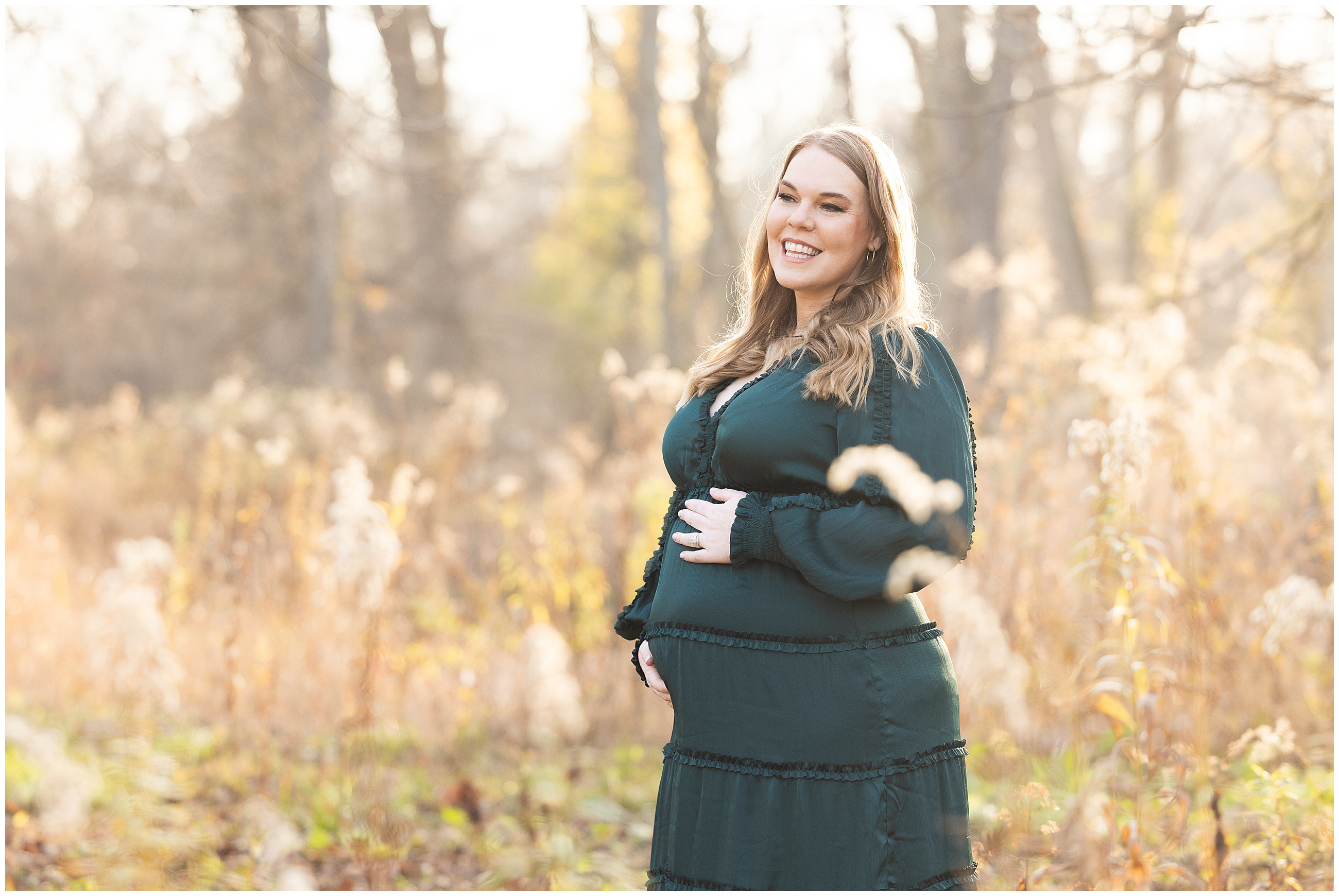 Pregnant woman in a dark green dress smiles in a sunlit autumn field, surrounded by soft-focus foliage and trees in the background.Columbus, Ohio maternity photographer.