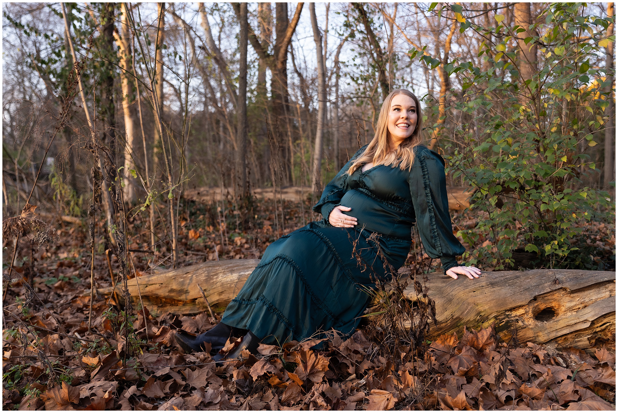 Pregnant woman in a dark green dress sits on a fallen log, smiling. Background is a wooded area with bare trees and fallen leaves.The golden sunset light is directly illuminating her.