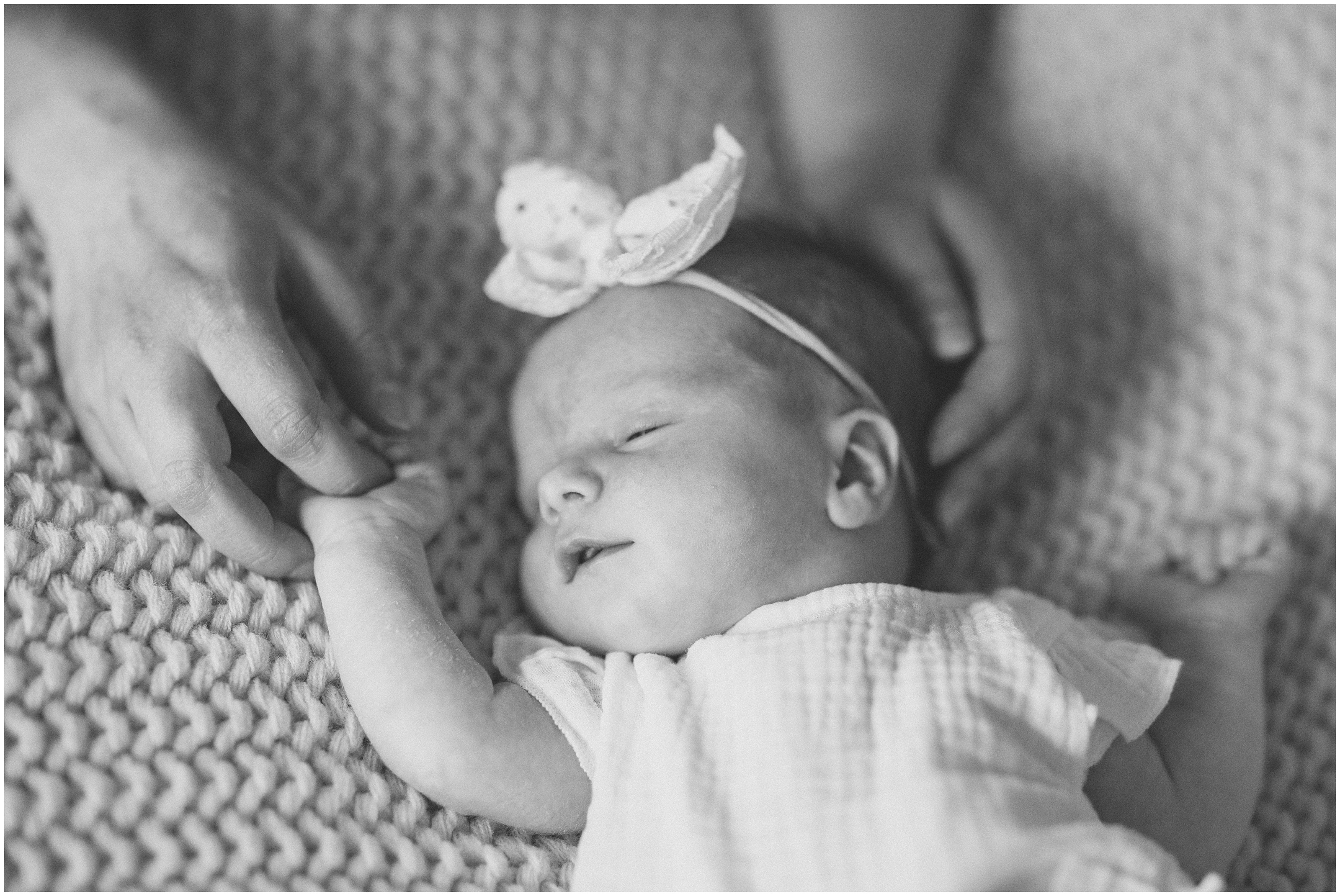 Black and white photo of sleeping newborn baby, cradled by adult hands on a knitted blanket, wearing a bow headband, Smiling family of four sitting on a bed, together, parents with toddler and newborn in a bright bedroom with cream pillows and bedding. Columbus Ohio newborn photography
