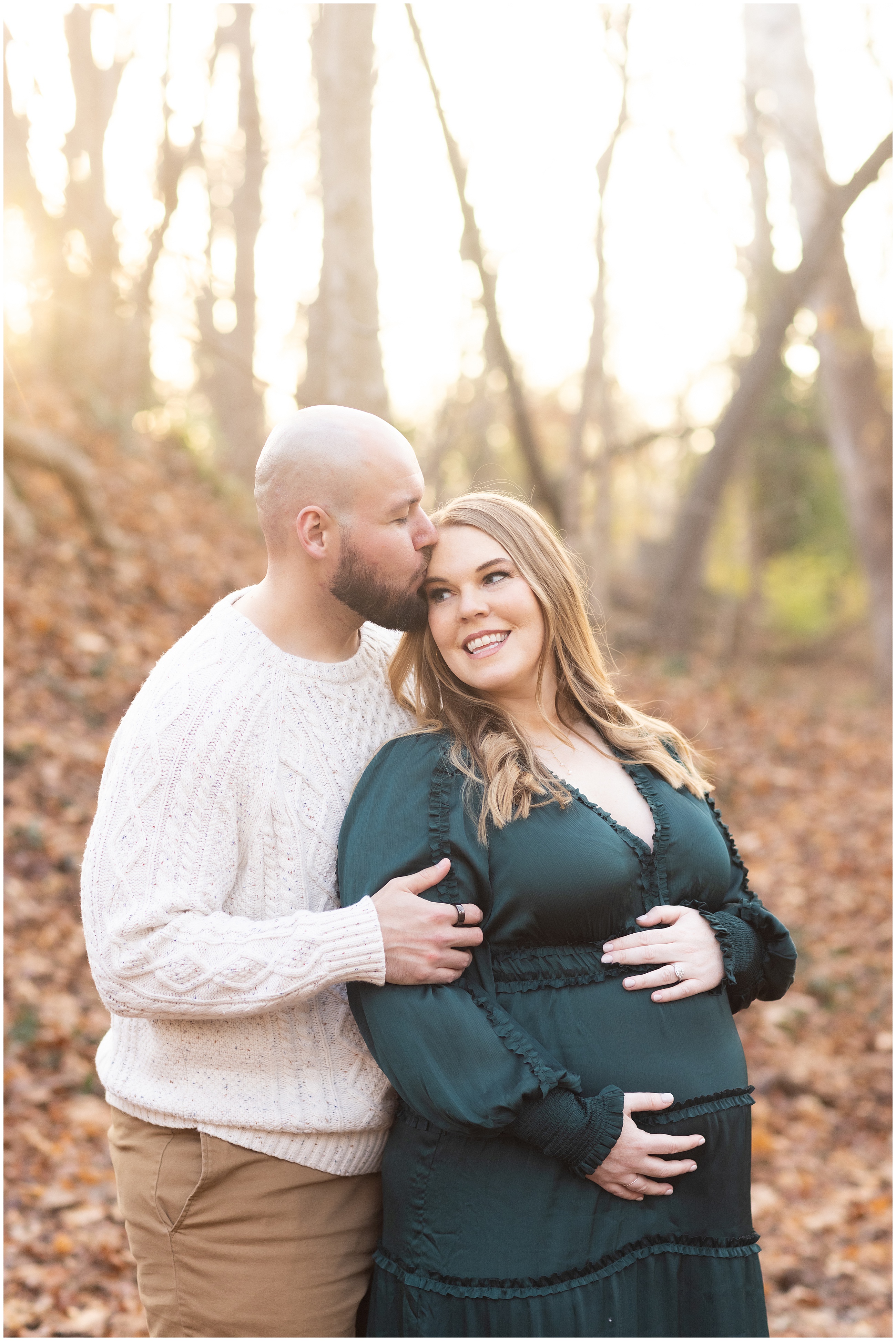 A couple embraces in a sunlit forest. The man kisses the woman's head as she smiles, holding her pregnant belly, wearing a green dress.