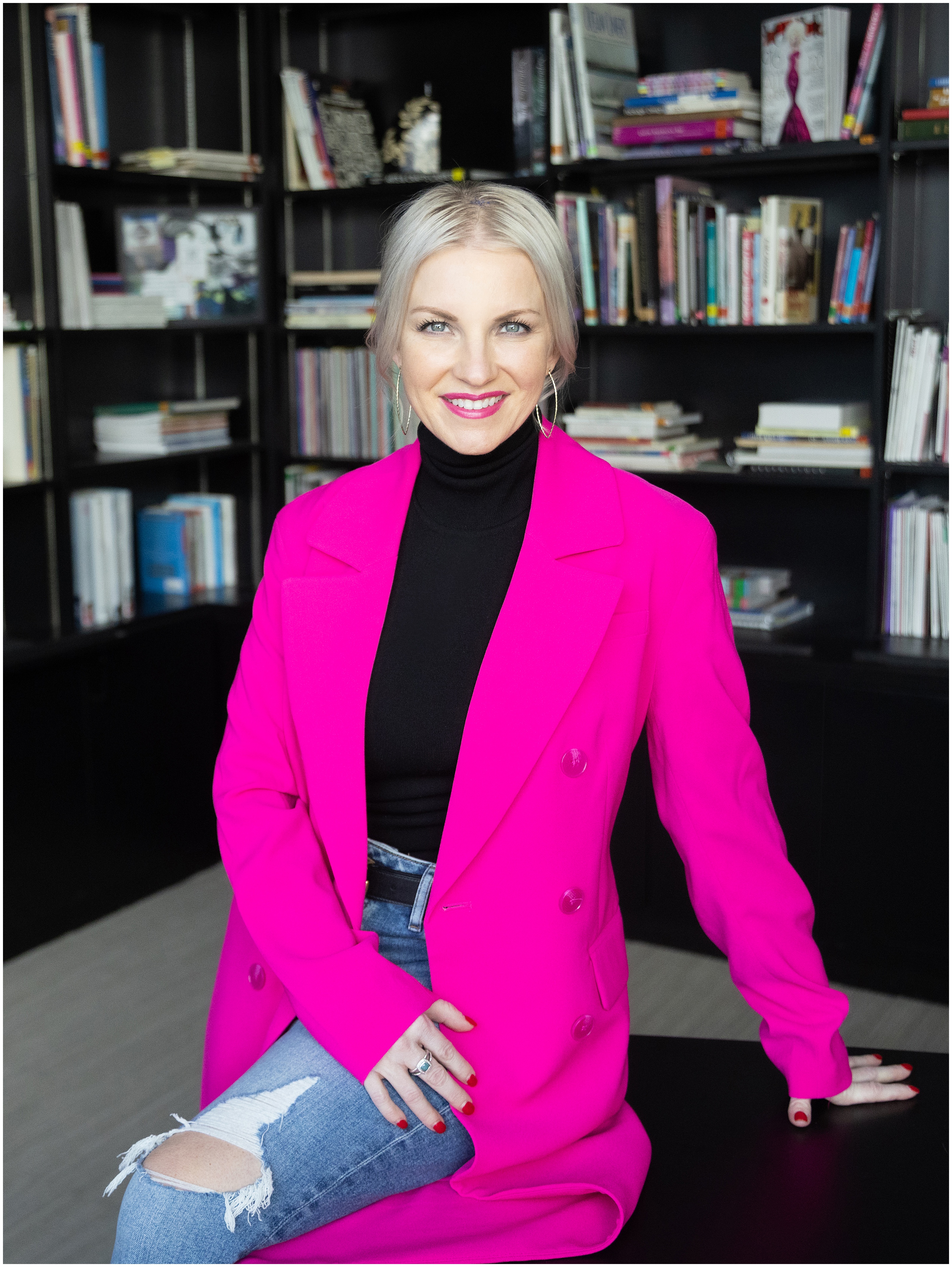 Stylish woman wearing a long bright pink blazer and ripped jeans sits in front of black bookshelves in an executive office.