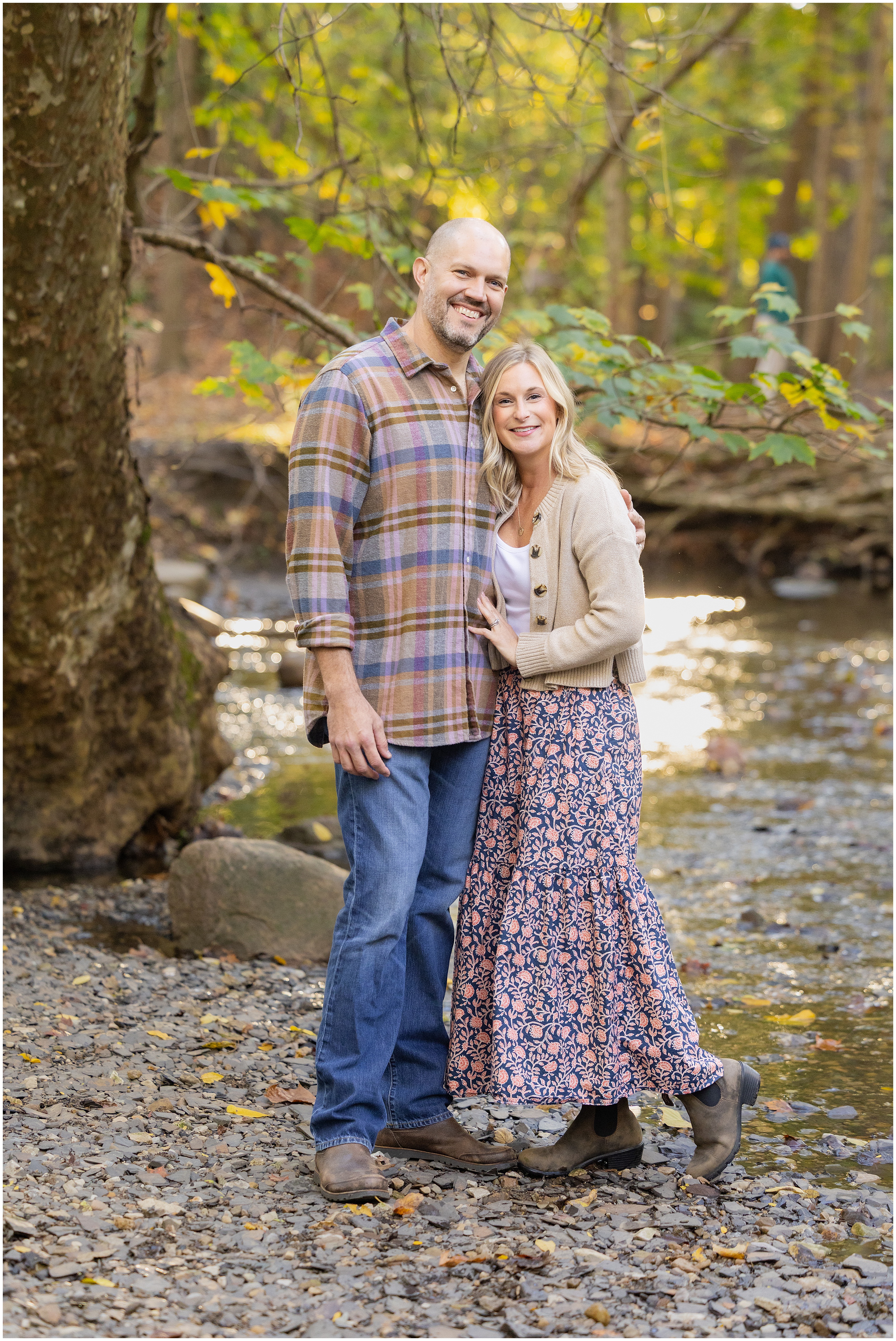 Smiling couple standing arm in arm by a creek in autumn woods, with golden leaves and a peaceful outdoor mood.
