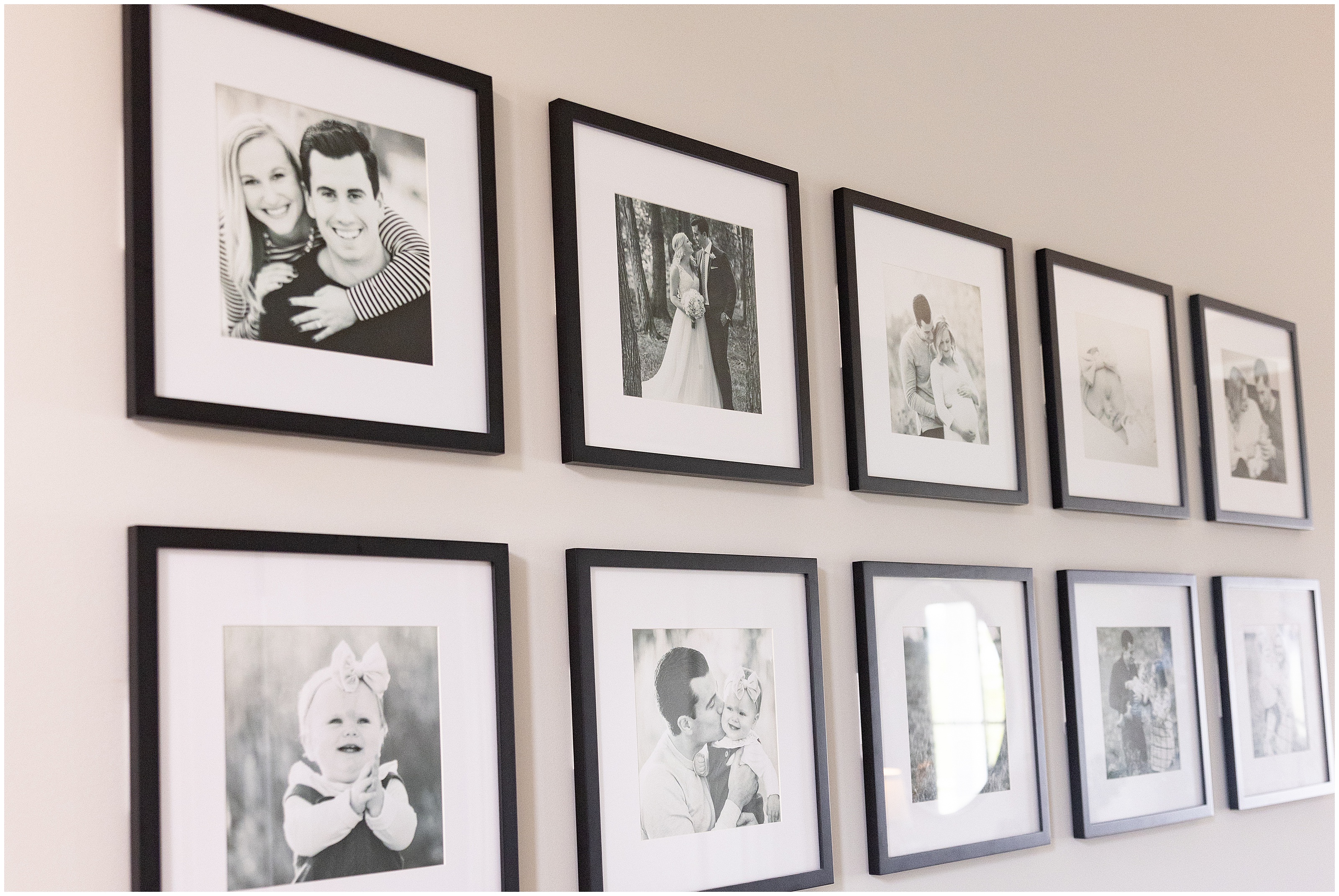 A modern gallery wall of black and white photographs in thin black frames showing couples, wedding and baby portraits, warm and sentimental