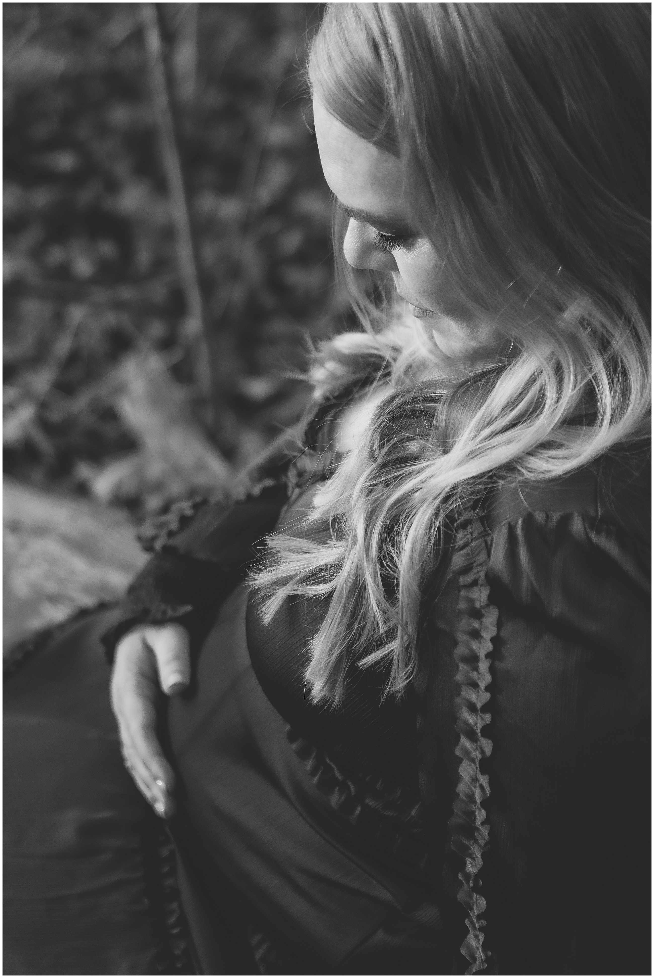 A pregnant woman in a dress sitting outdoors, hand on belly, with long blonde hair, creating a serene and contemplative mood. Black and white image.