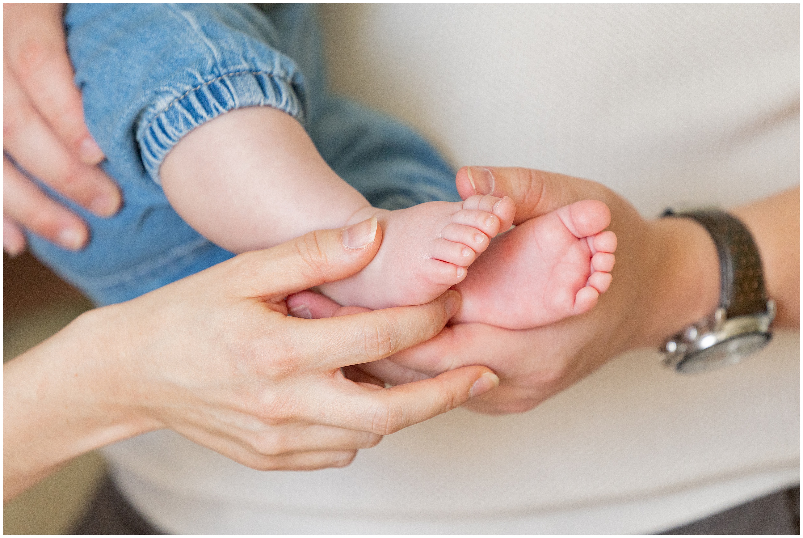 A close-up photo of a mother and father's hands gently holding a baby's feet. The baby wears blue pants. The background is soft and neutral, creating a tender, caring mood with warm lighting. Columbus Ohio newborn photography.