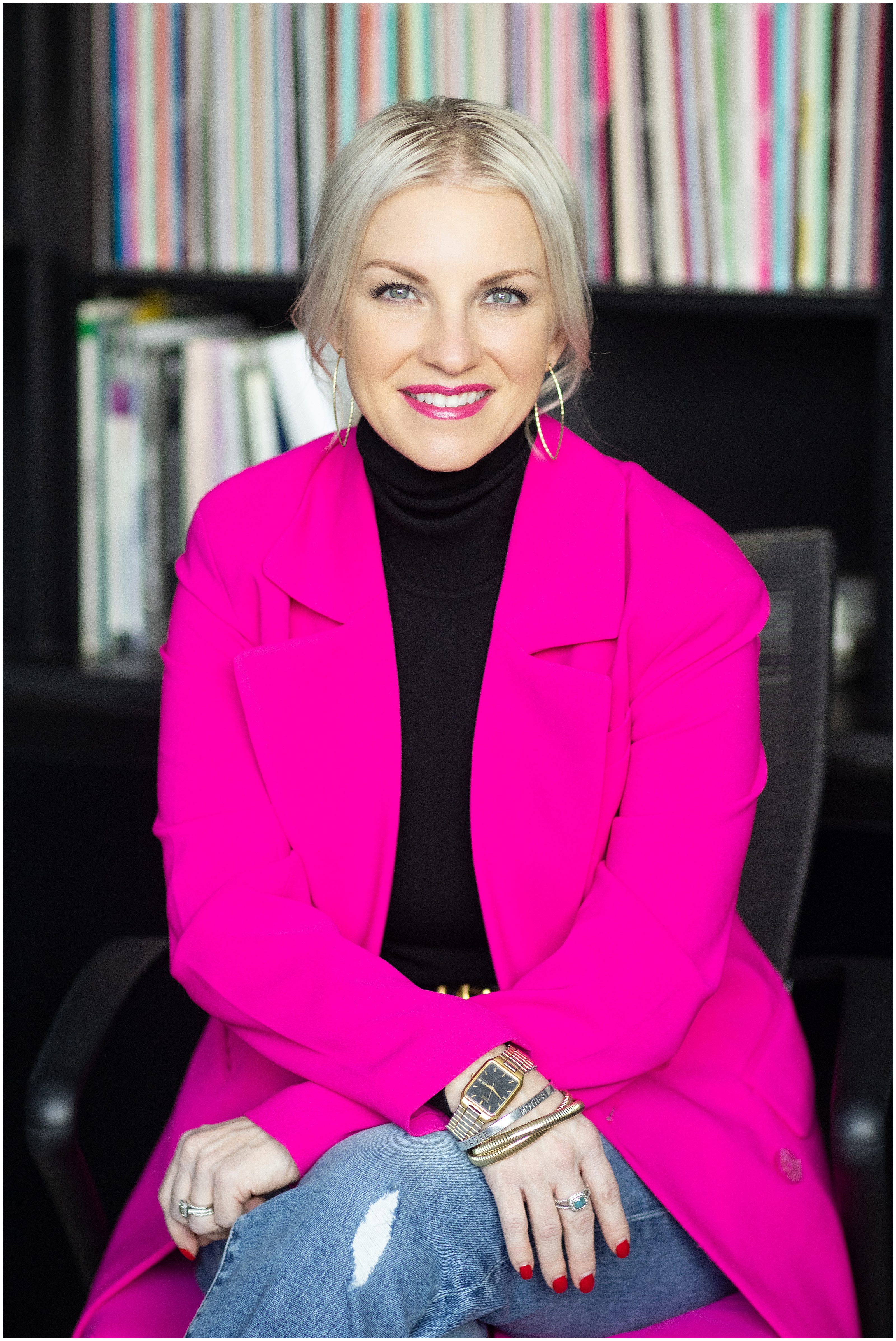 Smiling blonde woman in a bright pink blazer and black turtleneck sits in front of black bookshelves filled with colorful books in a modern office setting.