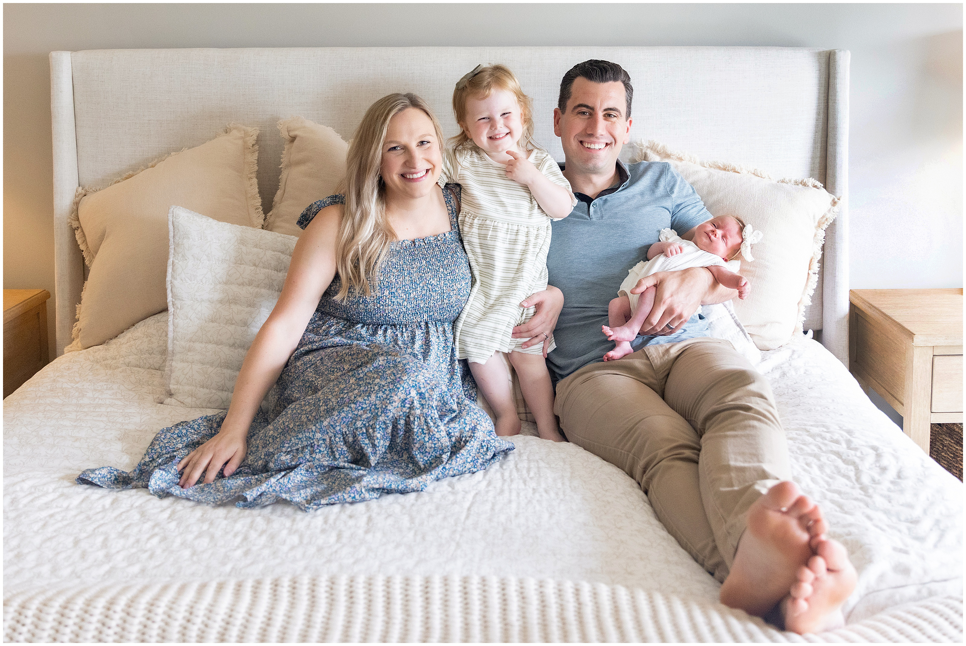 Smiling family of four sitting on a bed, together, parents with toddler and newborn in a bright bedroom with cream pillows and bedding. Columbus Ohio newborn photography
