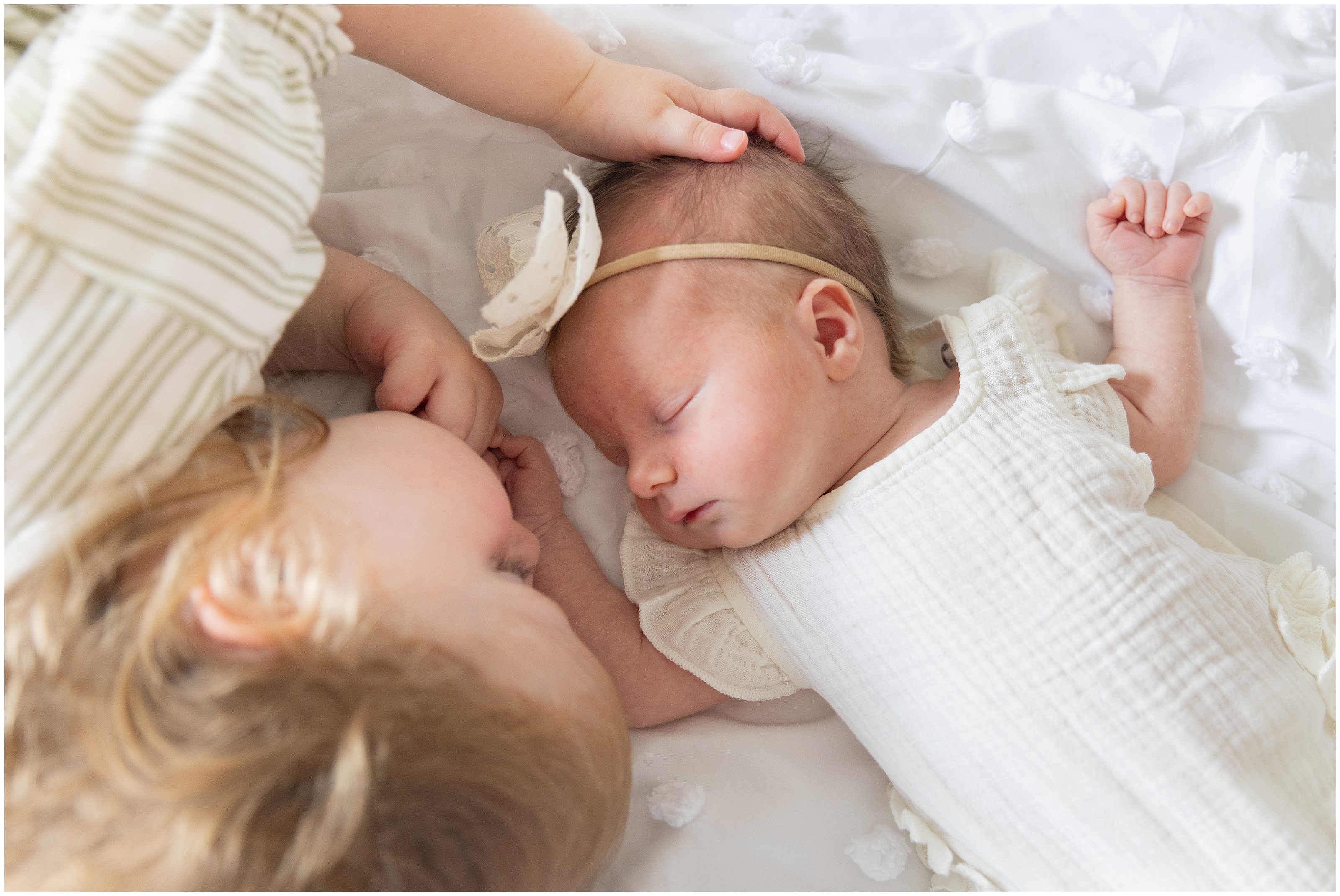 A sleeping newborn and toddler laying on soft white bedding facing each other, both wearing soft cream clothing, The toddler gently holds the baby's hand, very sweet moment, Columbus Ohio family photography