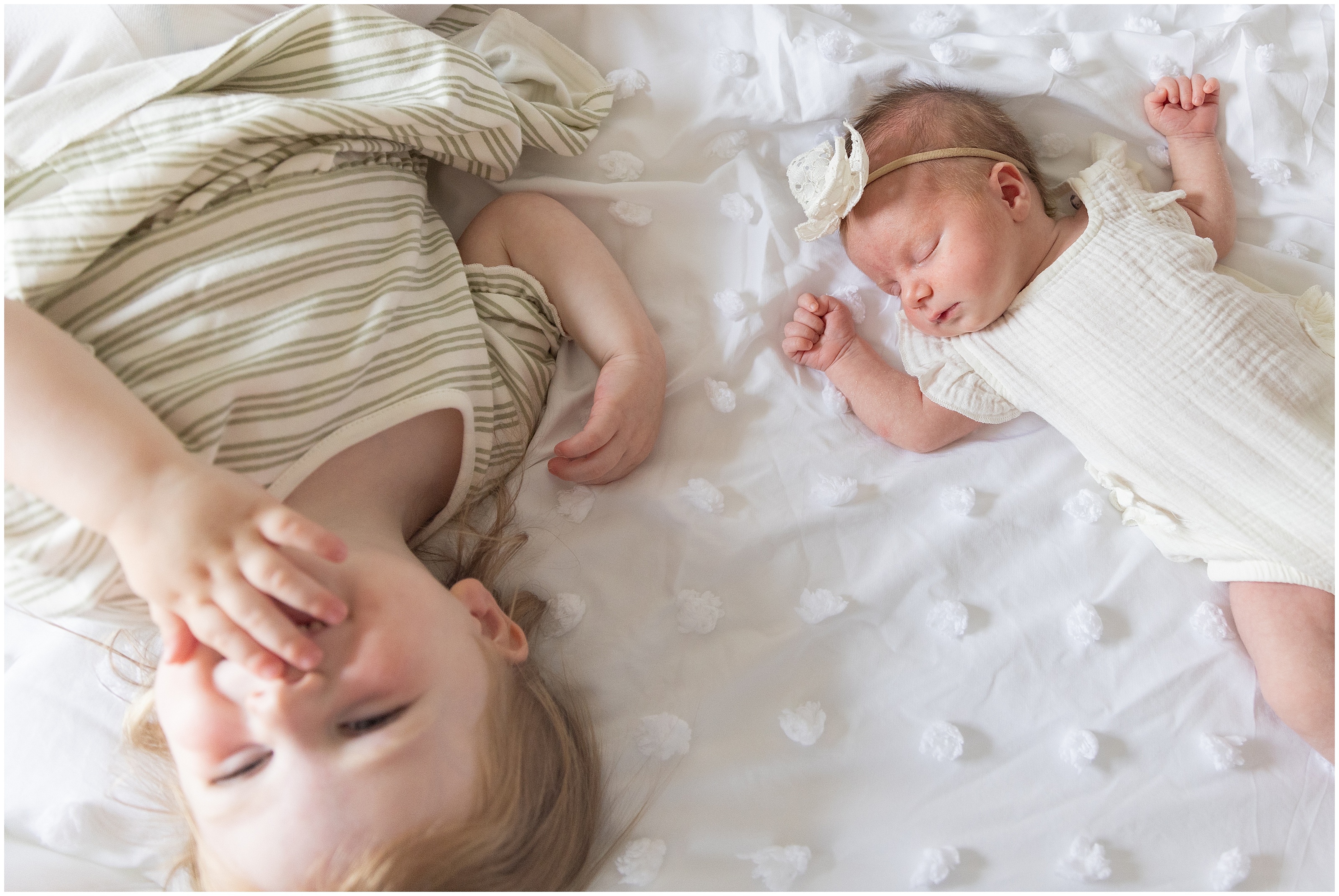 Giggling toddler and sleeping newborn on a white bed, both in cream outfits, with soft natural light