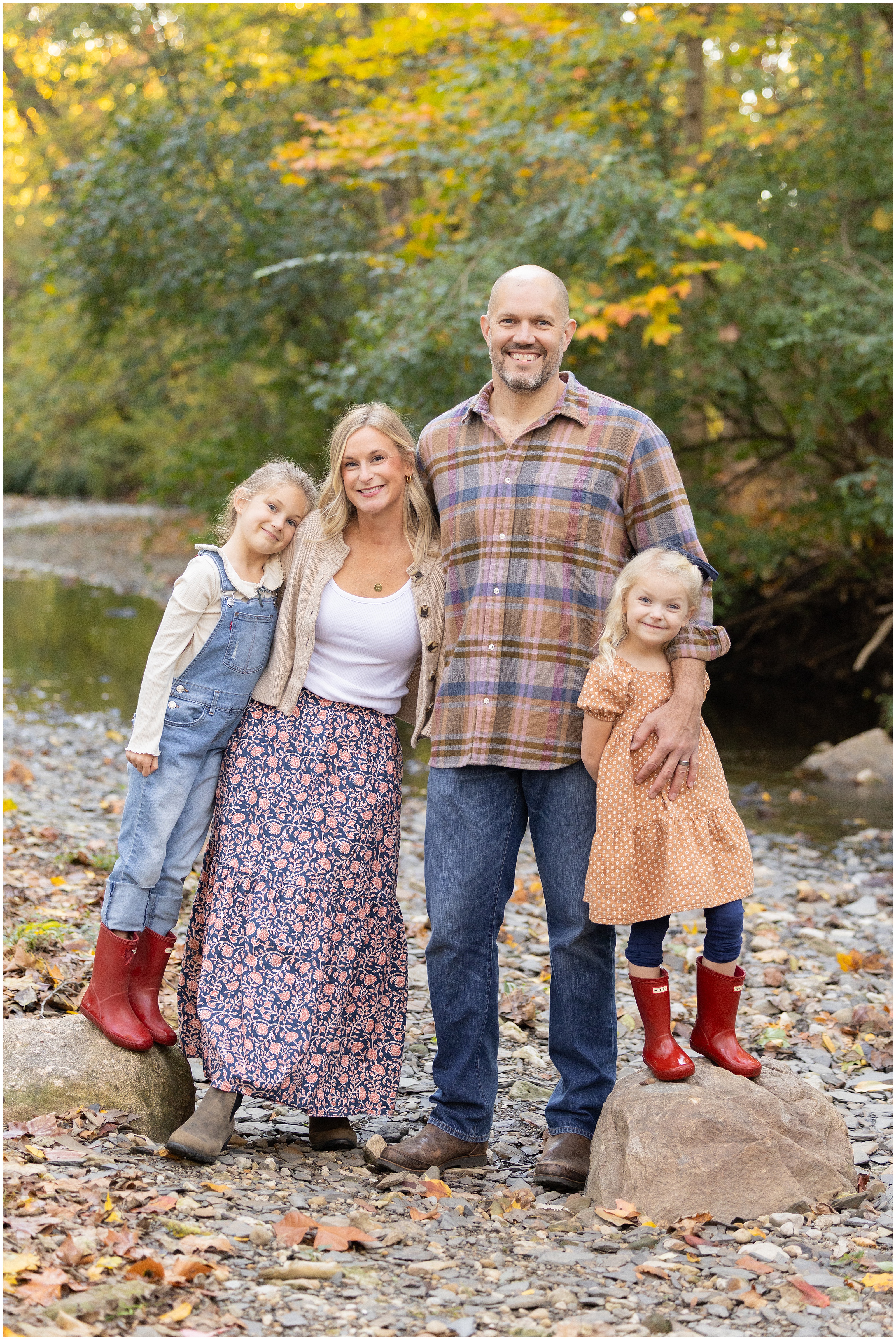 Family of four standing together in a wooded ravine setting in the fall, arms around each other smiling for a photo. There are two young girls in red rain boots, each standing on large rocks.
