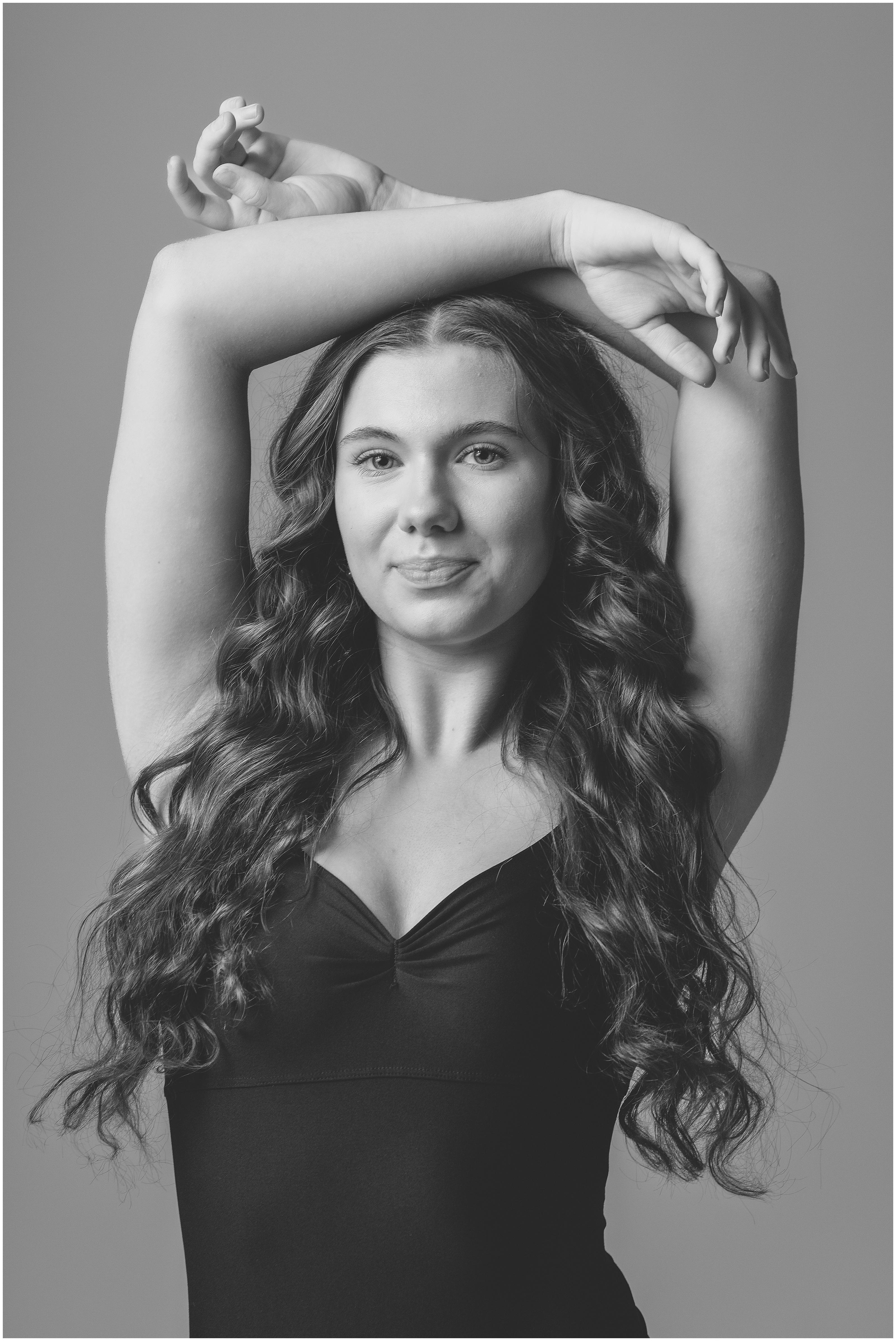 Black-and-white portrait of a young woman in a dark sleeveless tank top, arms raised overhead, posing calmly against a plain studio backdrop.