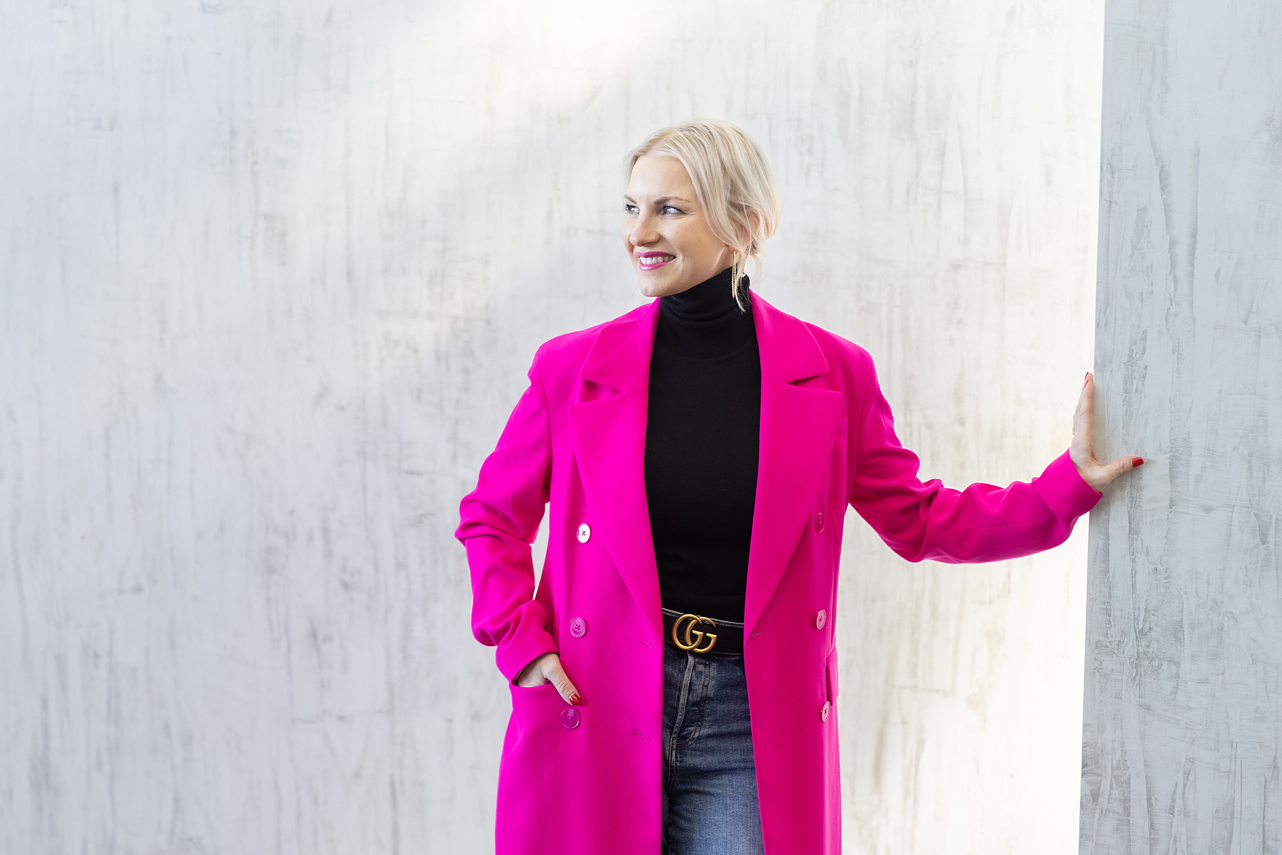 Woman wearing a long bright pink coat and black turtleneck leans on a white textured wall, posing confidently.