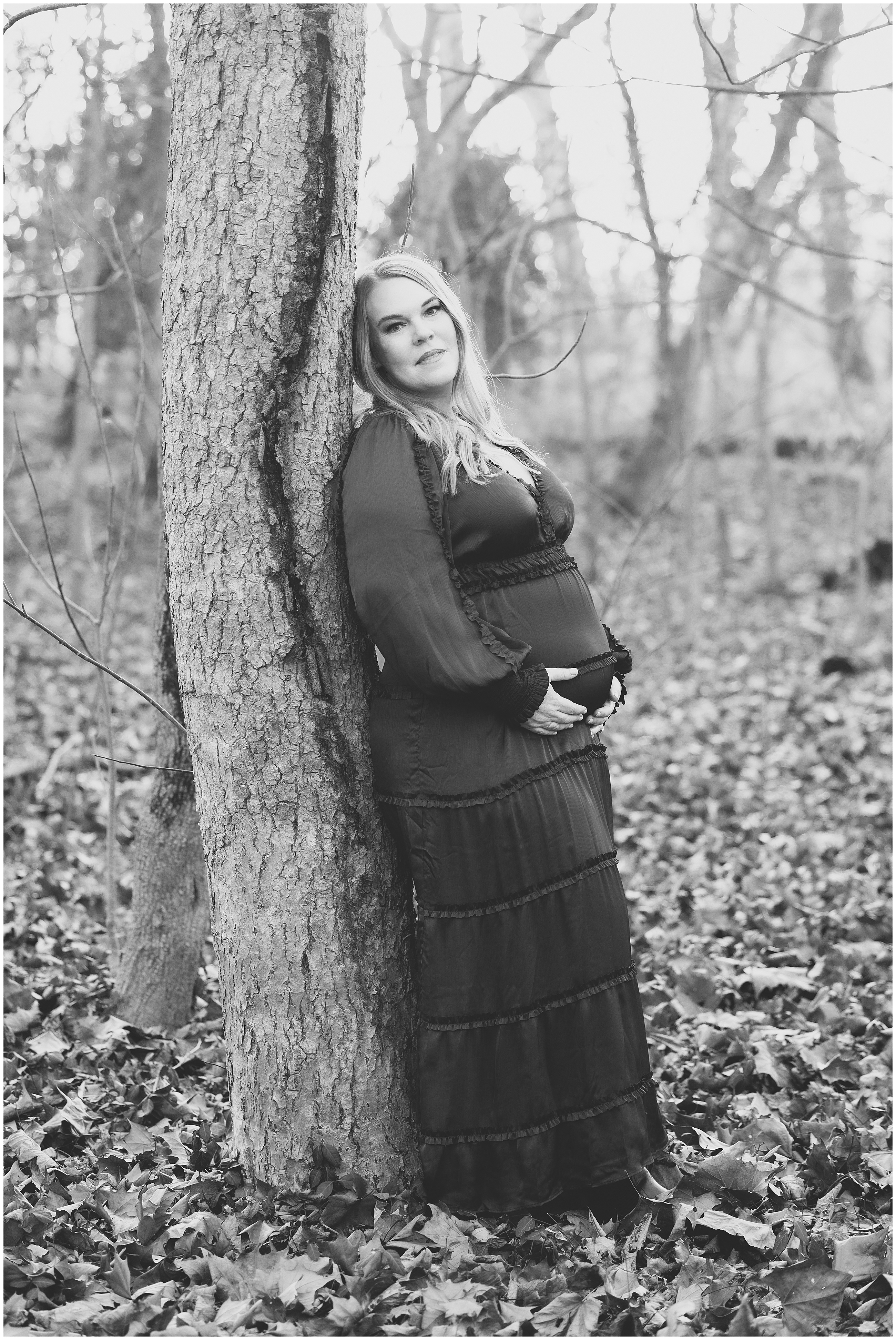 Pregnant woman in a long dress leans against a tree in a leafy forest. Black and white photo with a serene and contemplative mood. The ground is covered in leaves and most of the trees are bare.