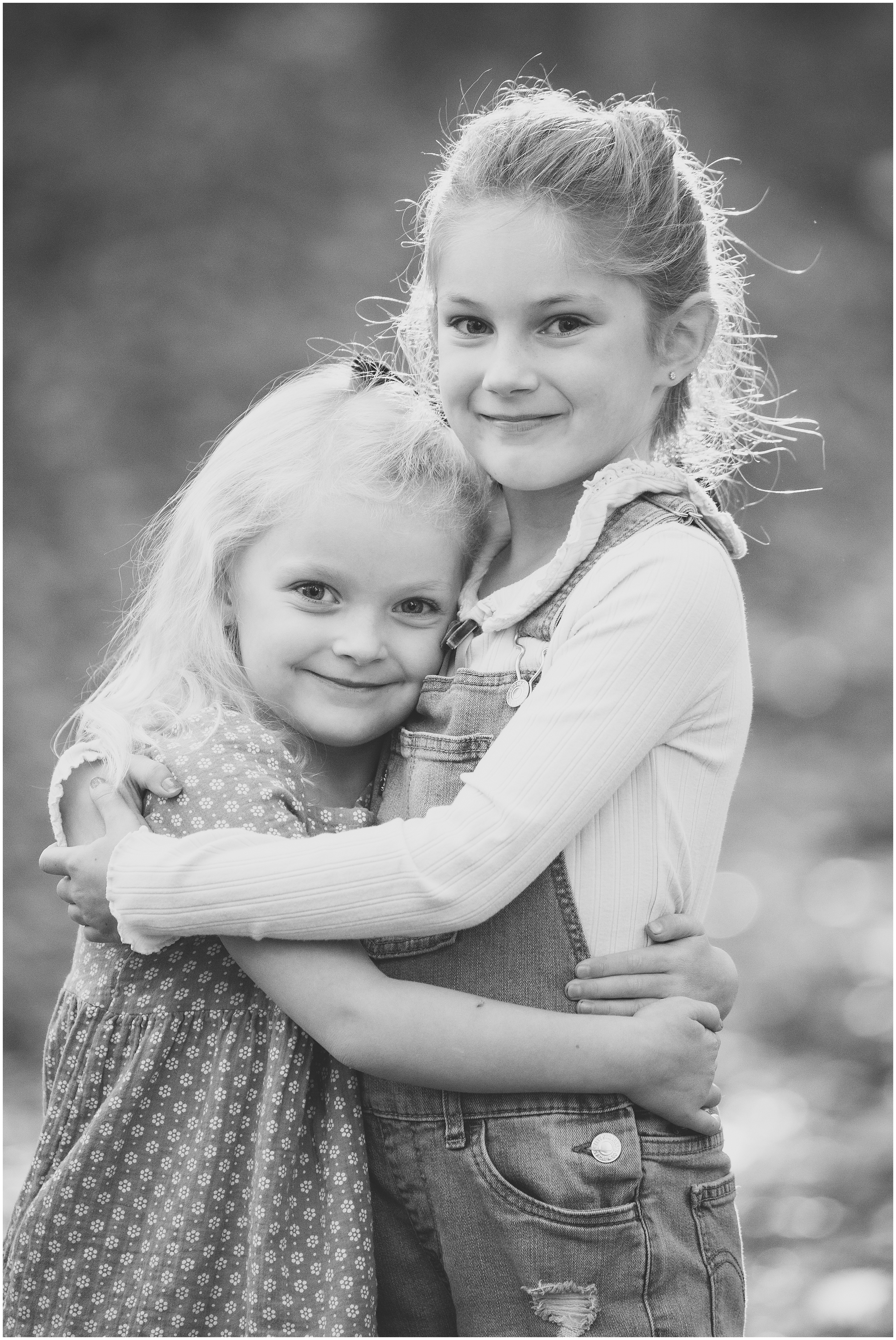 Two smiling young girls hug outdoors in a black-and-white portrait, one in overalls and one in a patterned dress.