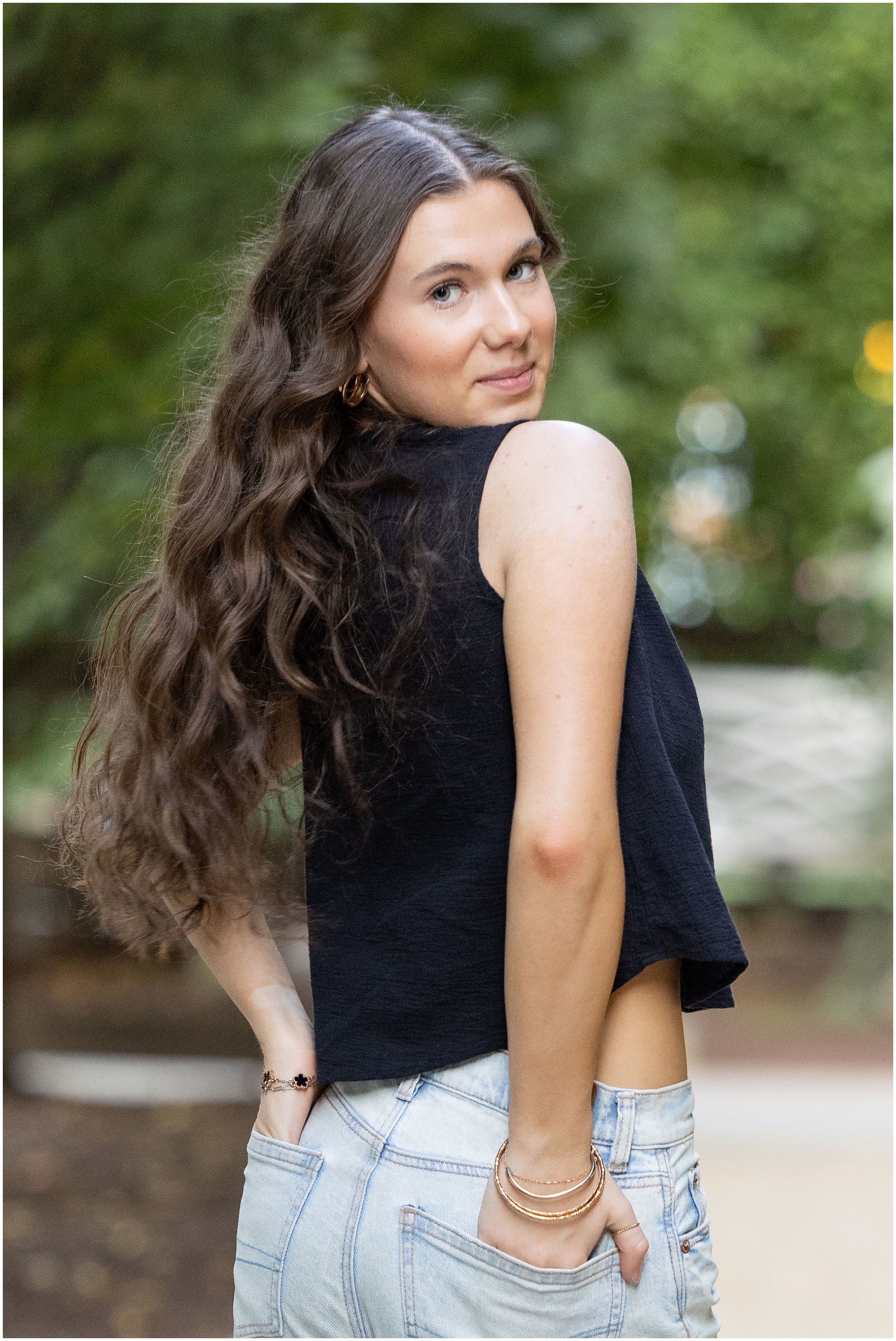 Young woman with long wavy hair looks back over her shoulder outdoors, wearing a black sleeveless top and light jeans. High school senior portrait