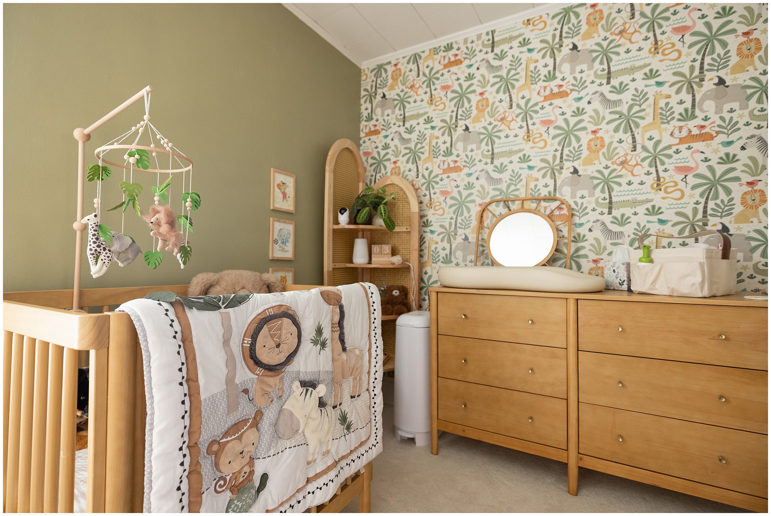 A nursery with a crib featuring animal-patterned bedding, a hanging mobile, and jungle-themed wallpaper. A wooden dresser with decor.