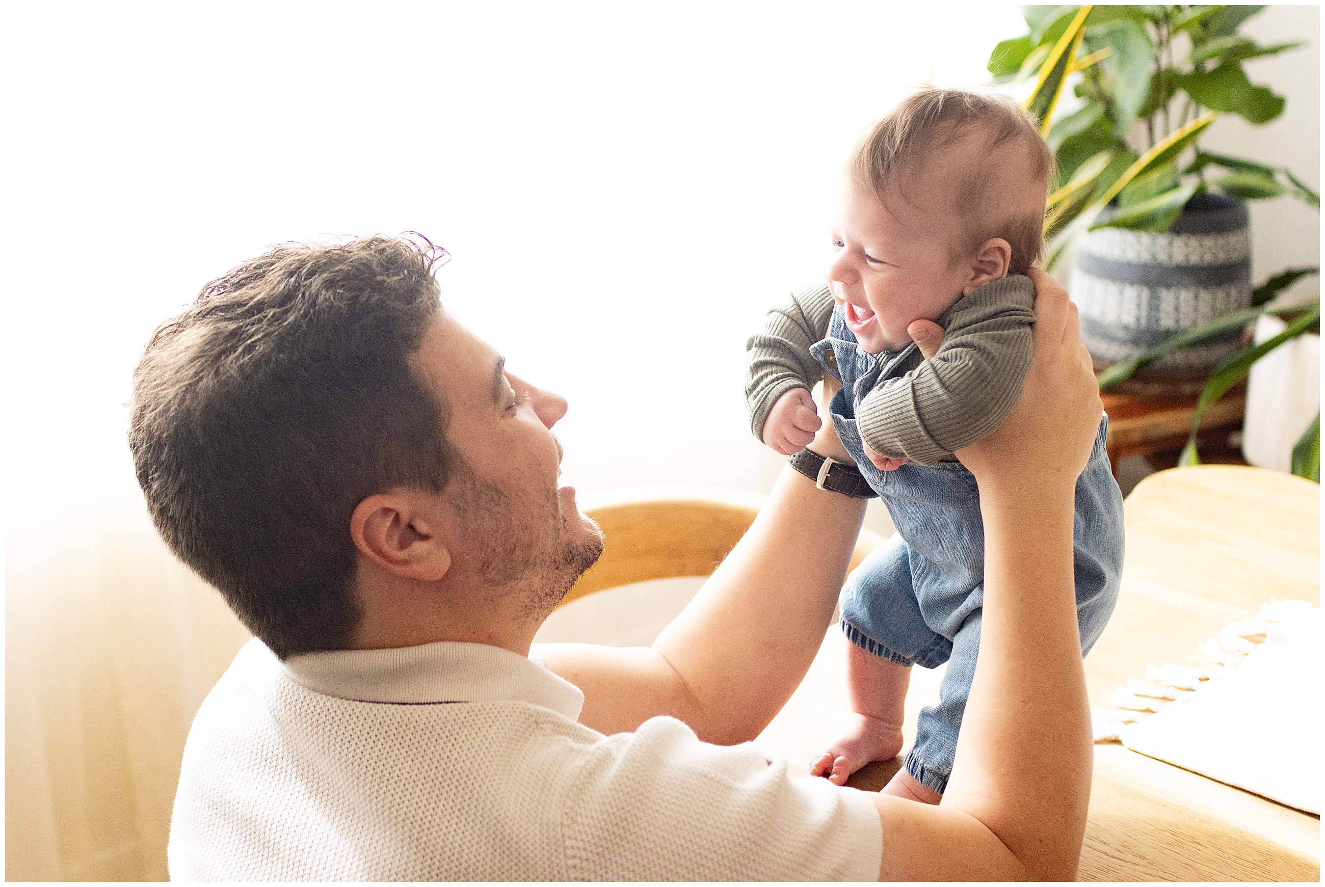 A smiling man lifts a laughing baby indoors by a plant. Baby wears denim and a green sweater. Soft lighting creates a warm atmosphere. Columbus Ohio newborn photographer.