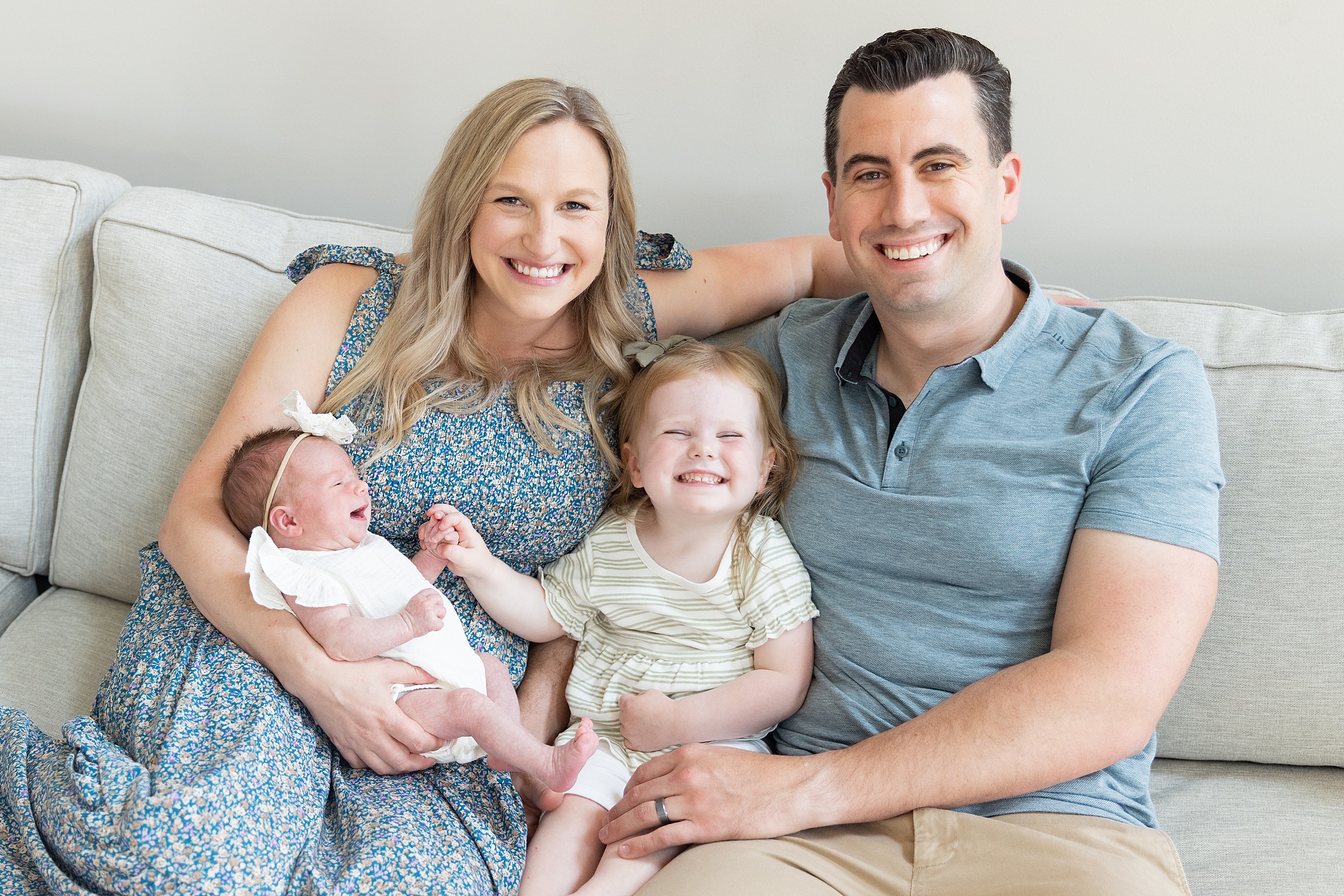 Smiling family of four sits on a light couch, parents holding a newborn and toddler in a bright living room. Family photography