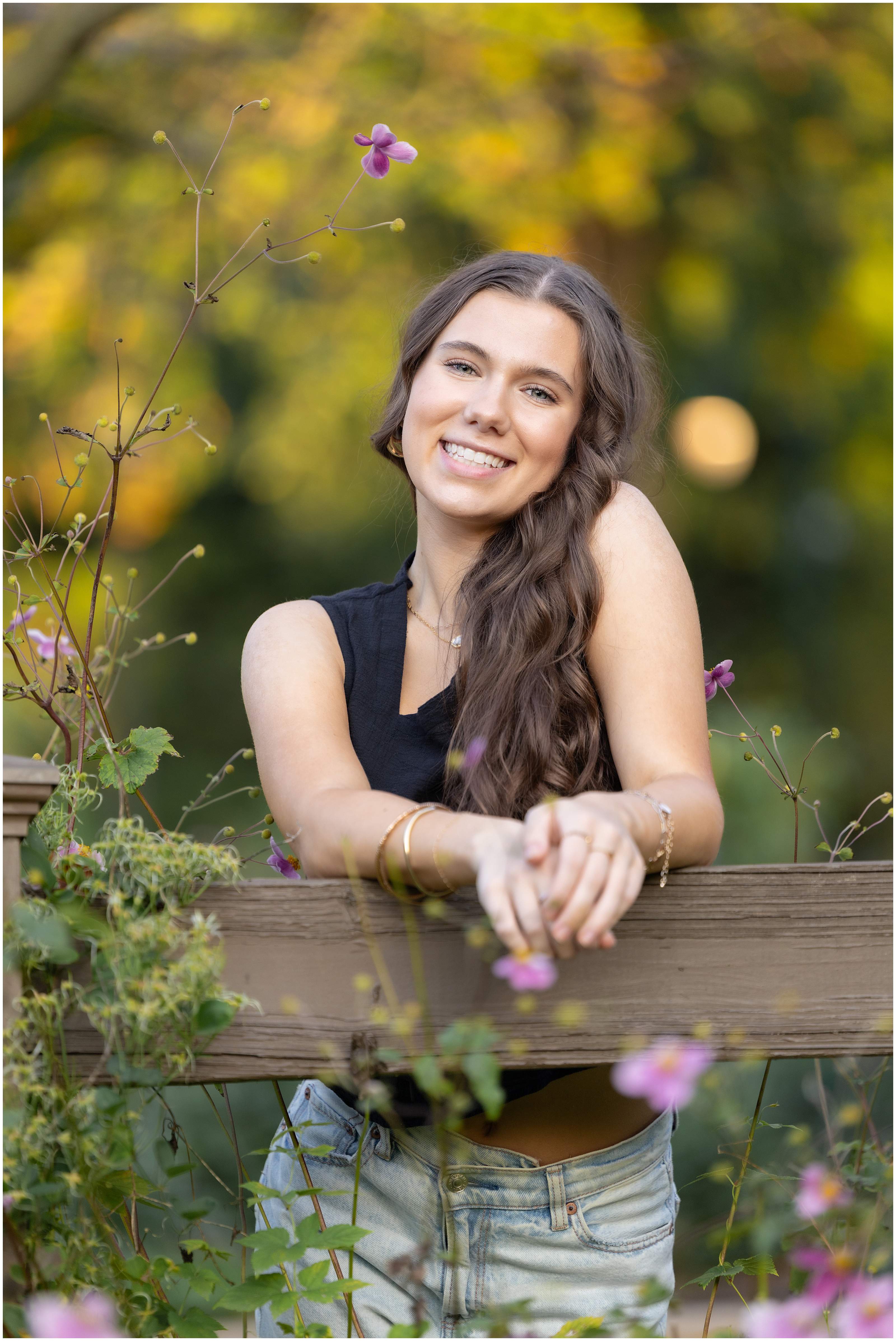 Smiling young woman with long brown hair leans on a wooden fence in a flower garden, with blurred yellow-green background and pink wildflower blooms.