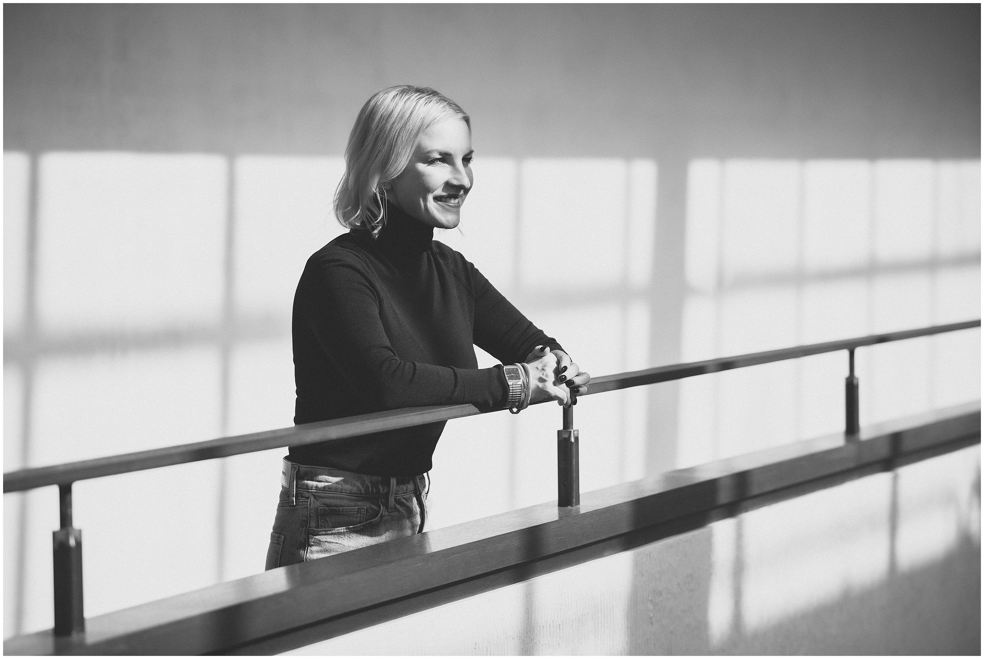 Dramatic black and white image of a smiling woman in a black turtleneck leaning on a railing in the hallway of a modern office building. The shadows from the window panes create a bold pattern over the image.