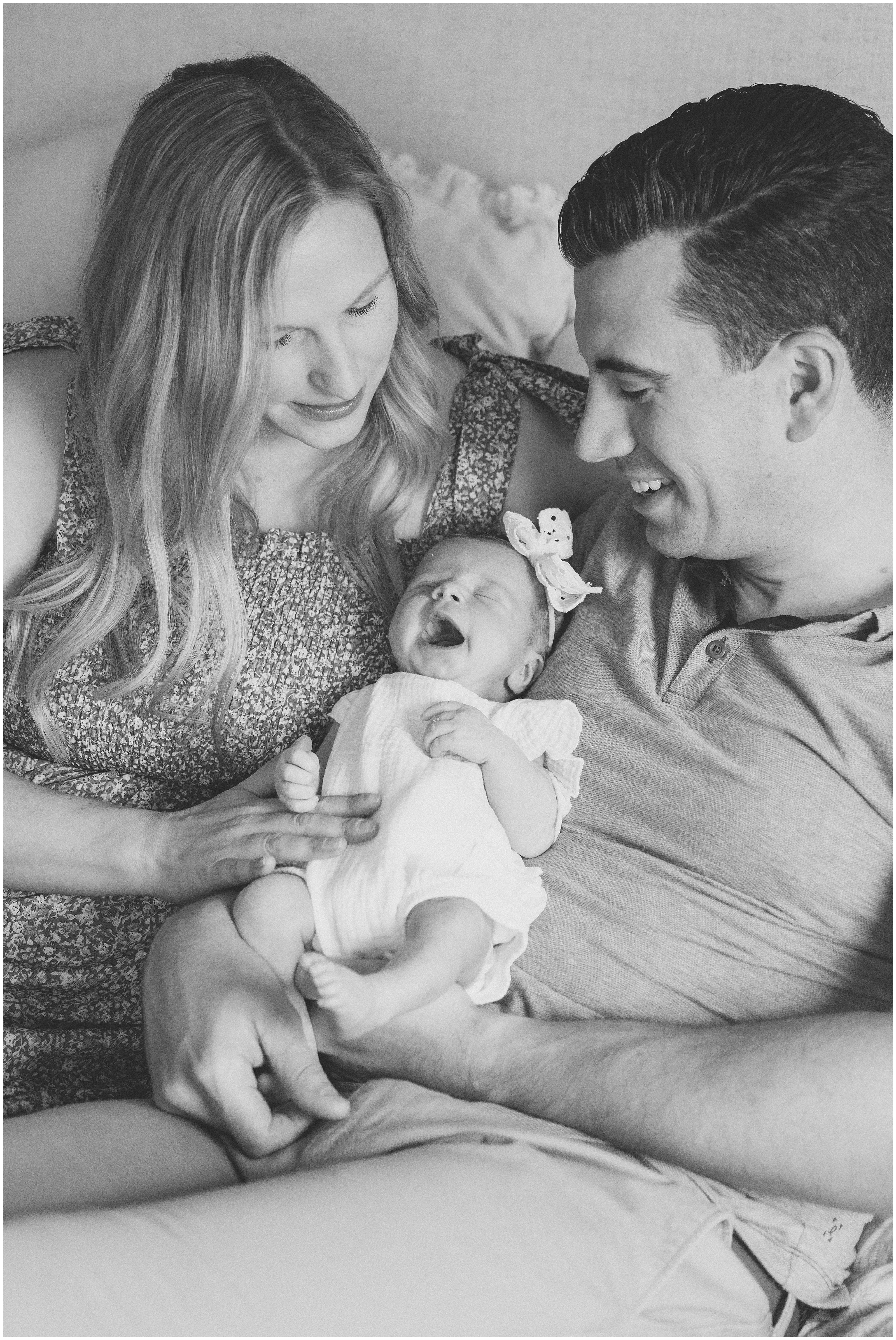 Black-and-white photo of smiling parents cradling a yawning newborn with a bow headband on a bed, sharing a tender moment Smiling family of four sitting on a bed, together, parents with toddler and newborn in a bright bedroom with cream pillows and bedding. Columbus Ohio newborn photography