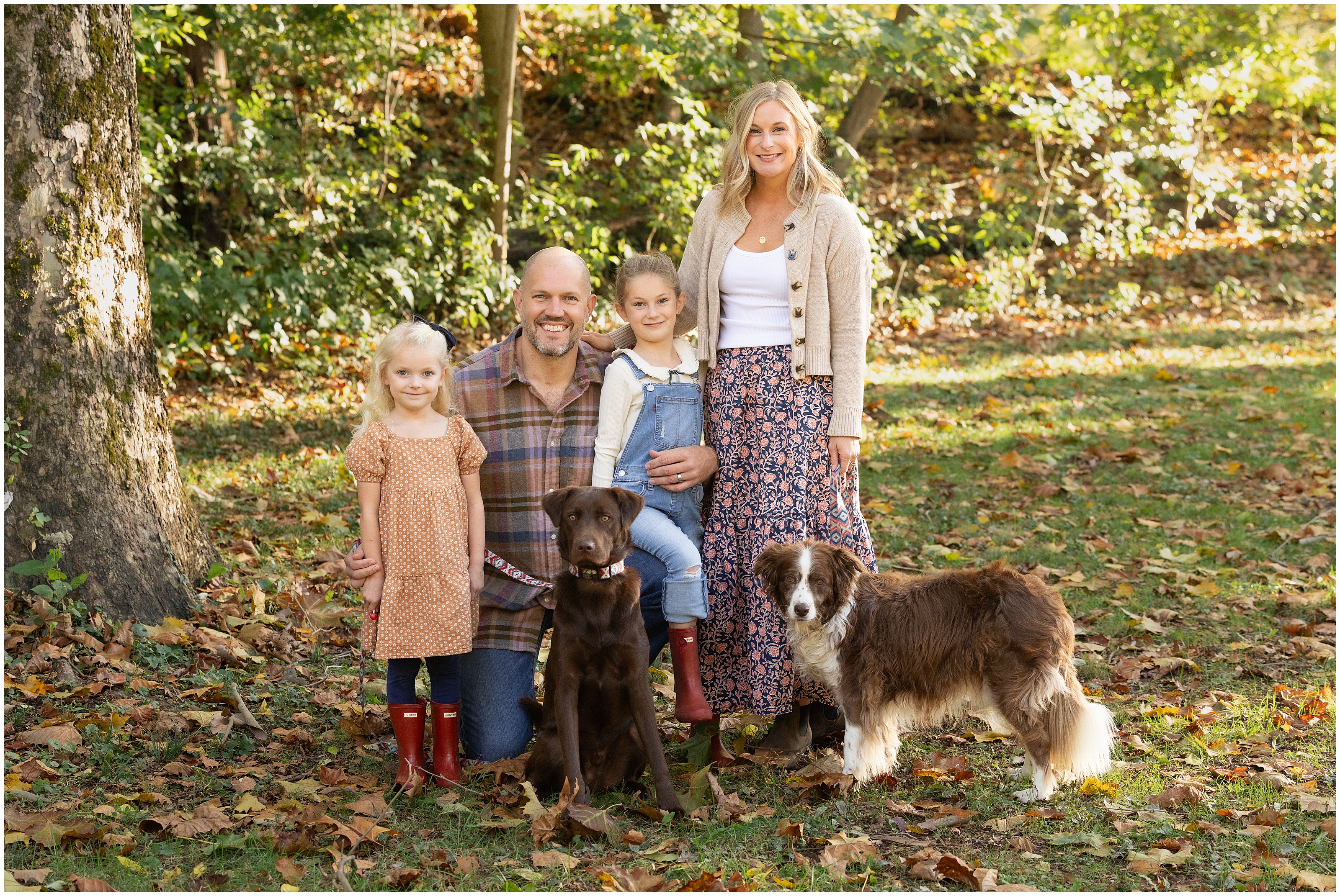 Smiling family of four with two dogs posing in a leafy autumn yard, with warm sunlight and casual outfits. Family photography in Columbus, Ohio