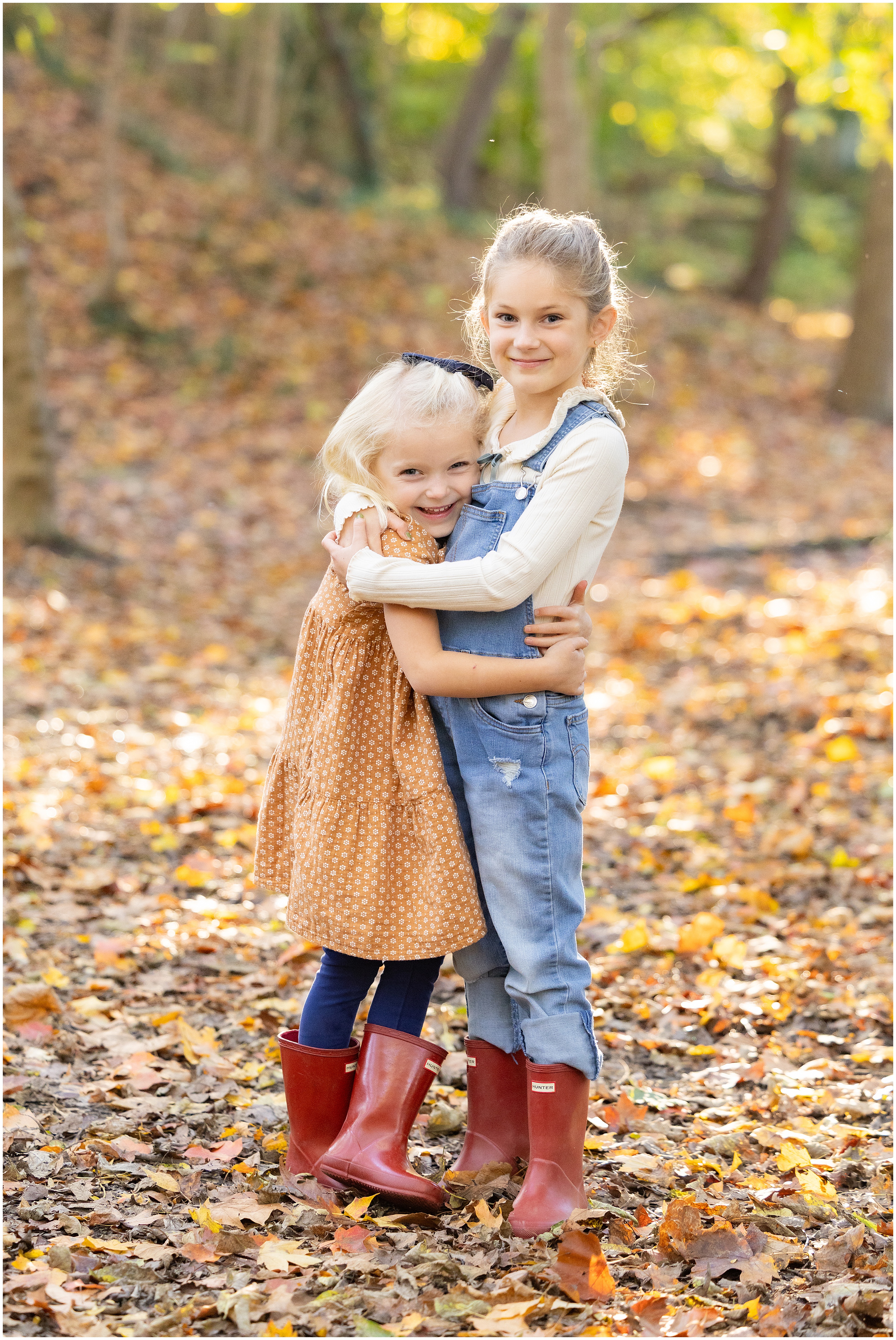 Two young girls hugging  and smiling at the camera, wearing red rain boots, denim overalls and a dress, a colorful fall setting in the woods.