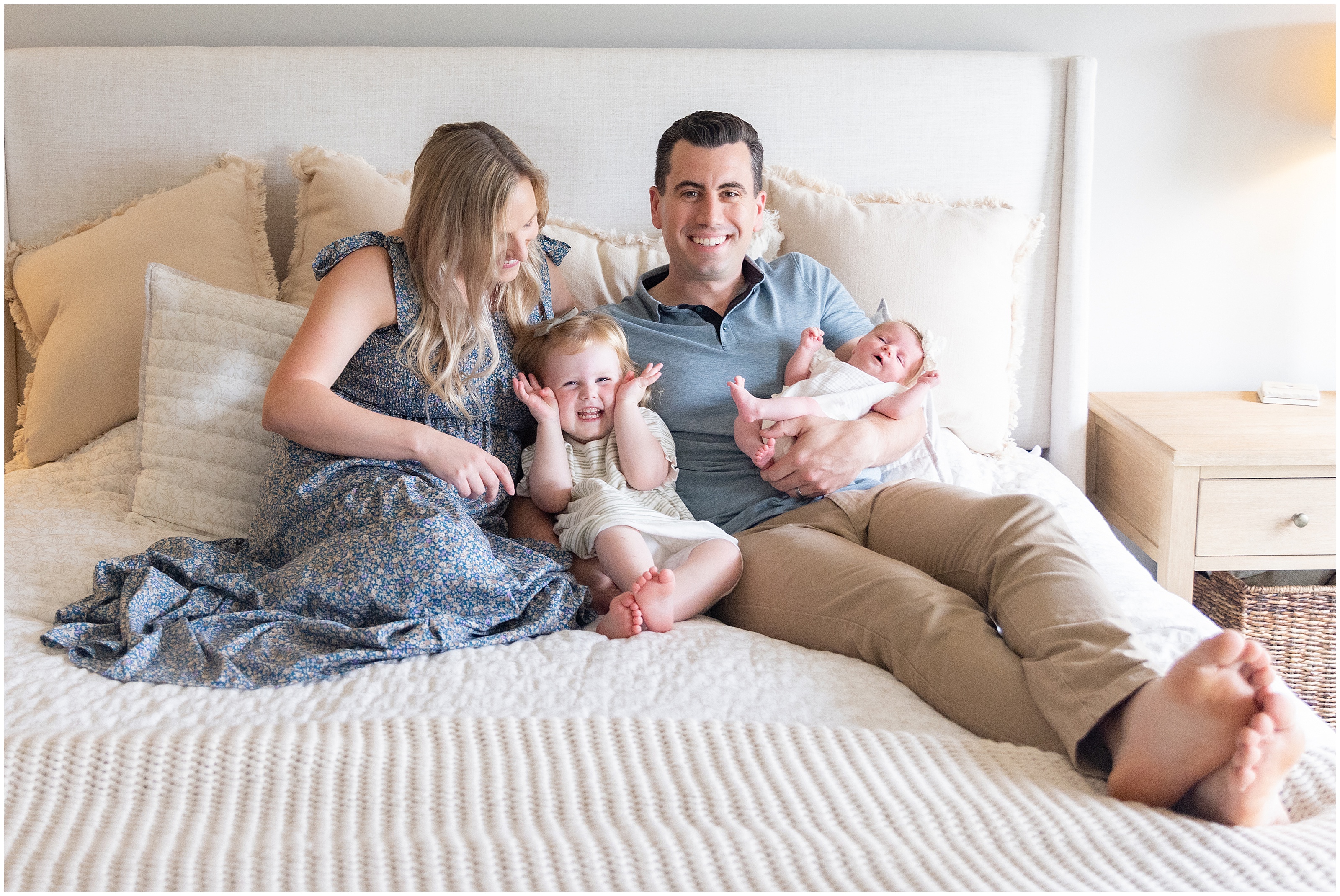Smiling family of four sitting on a bed, together, parents with toddler and newborn in a bright bedroom with cream pillows and bedding. Columbus Ohio newborn photography